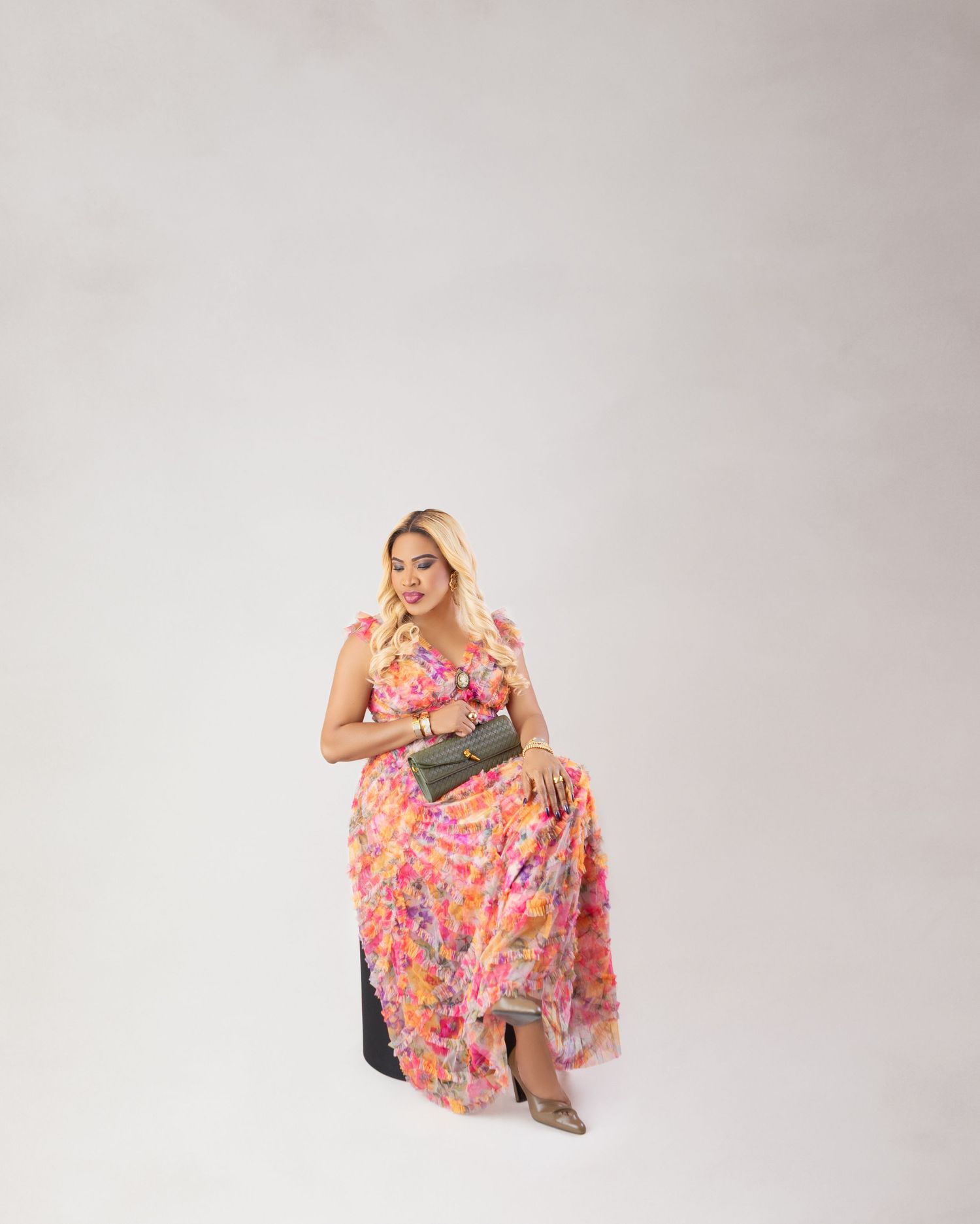 Woman in pink floral maxi dress holding olive green clutch bag, seated against white background.