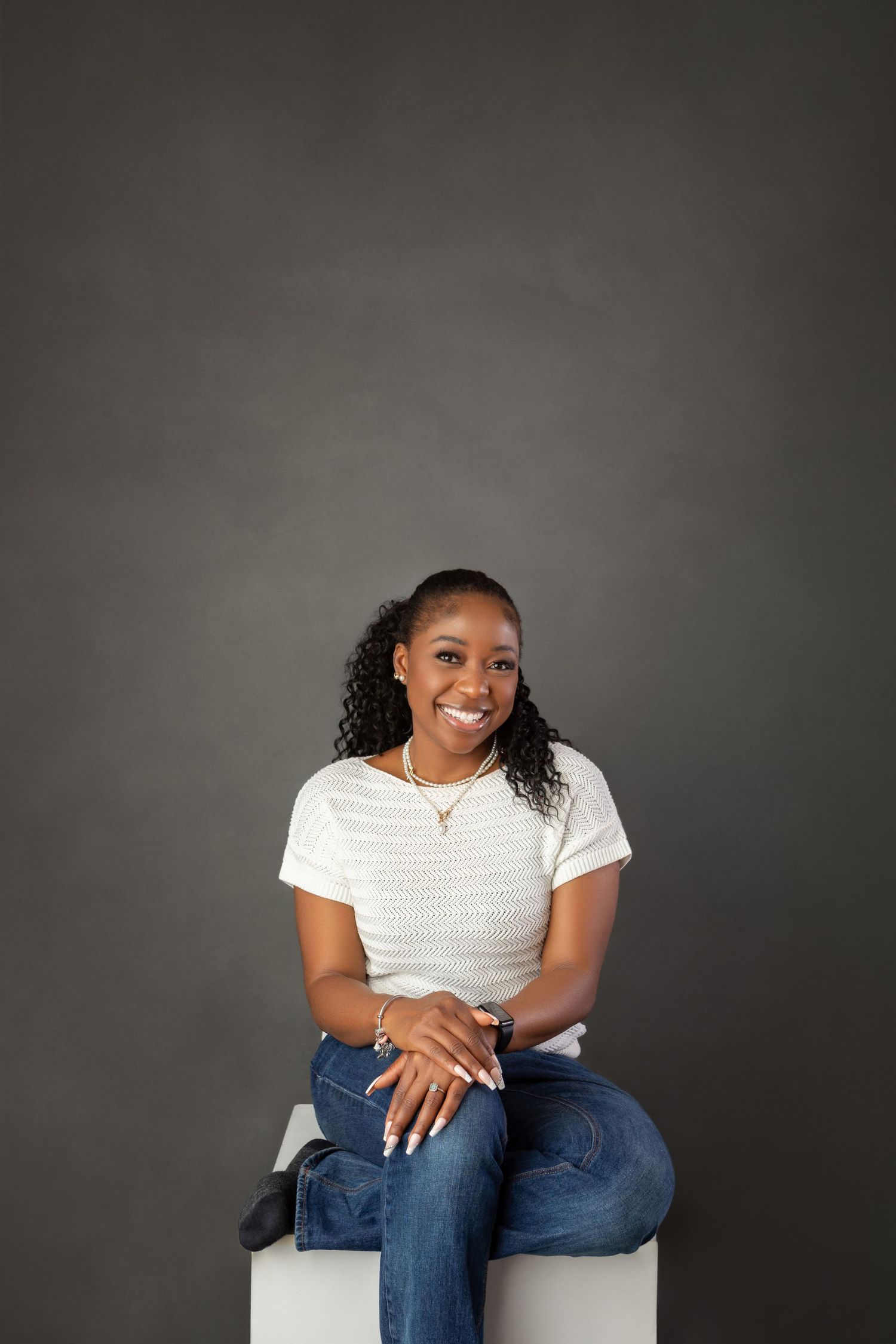 Smiling young Black woman in white top and jeans sitting on white cube against gray studio background.