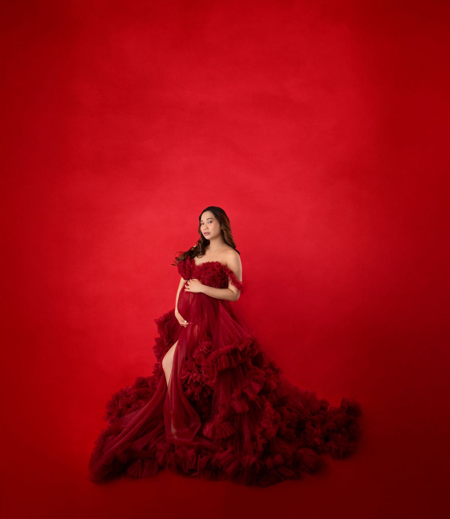 A pregnant woman in a dramatic dark red off-shoulder tulle gown poses against a bold red background.