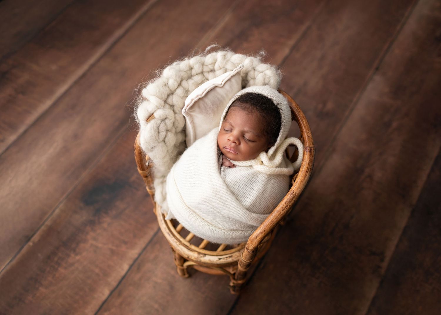 Newborn baby wrapped in cream swaddle sleeping in a small wicker bassinet on wood floor.