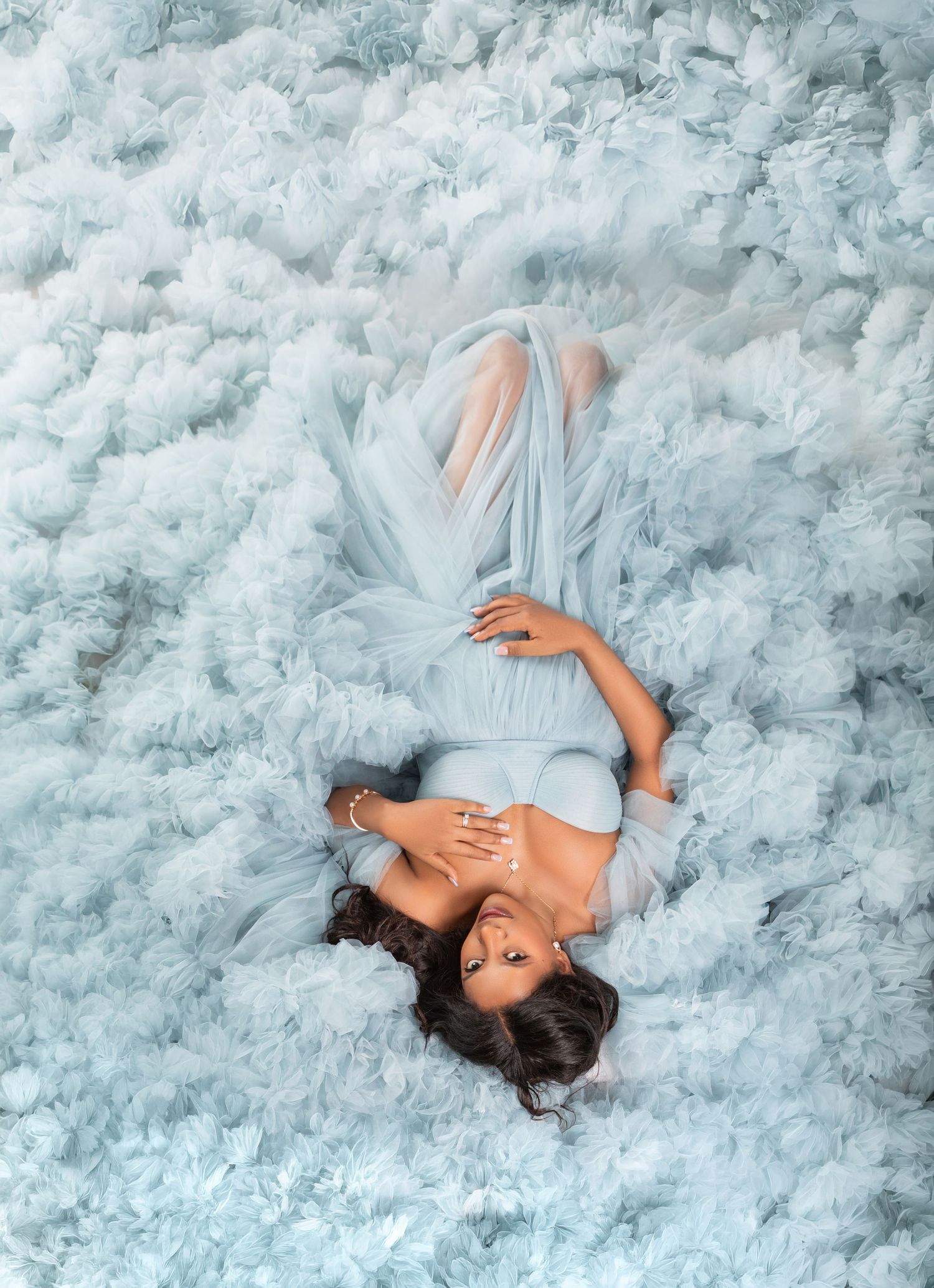 Pregnant woman in light blue tulle gown lying among soft blue clouds in a dreamy maternity photo.