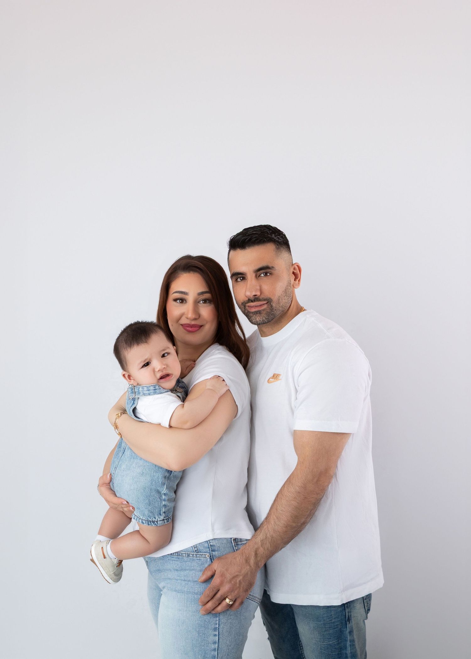Happy family of three in white tops and jeans posing with baby in a bright studio.
