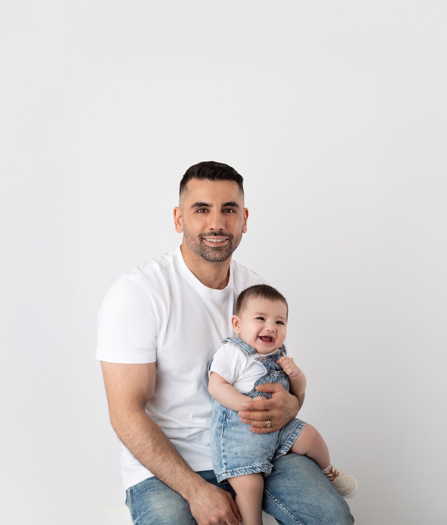Father in white t-shirt holding laughing baby in denim overalls on white background.