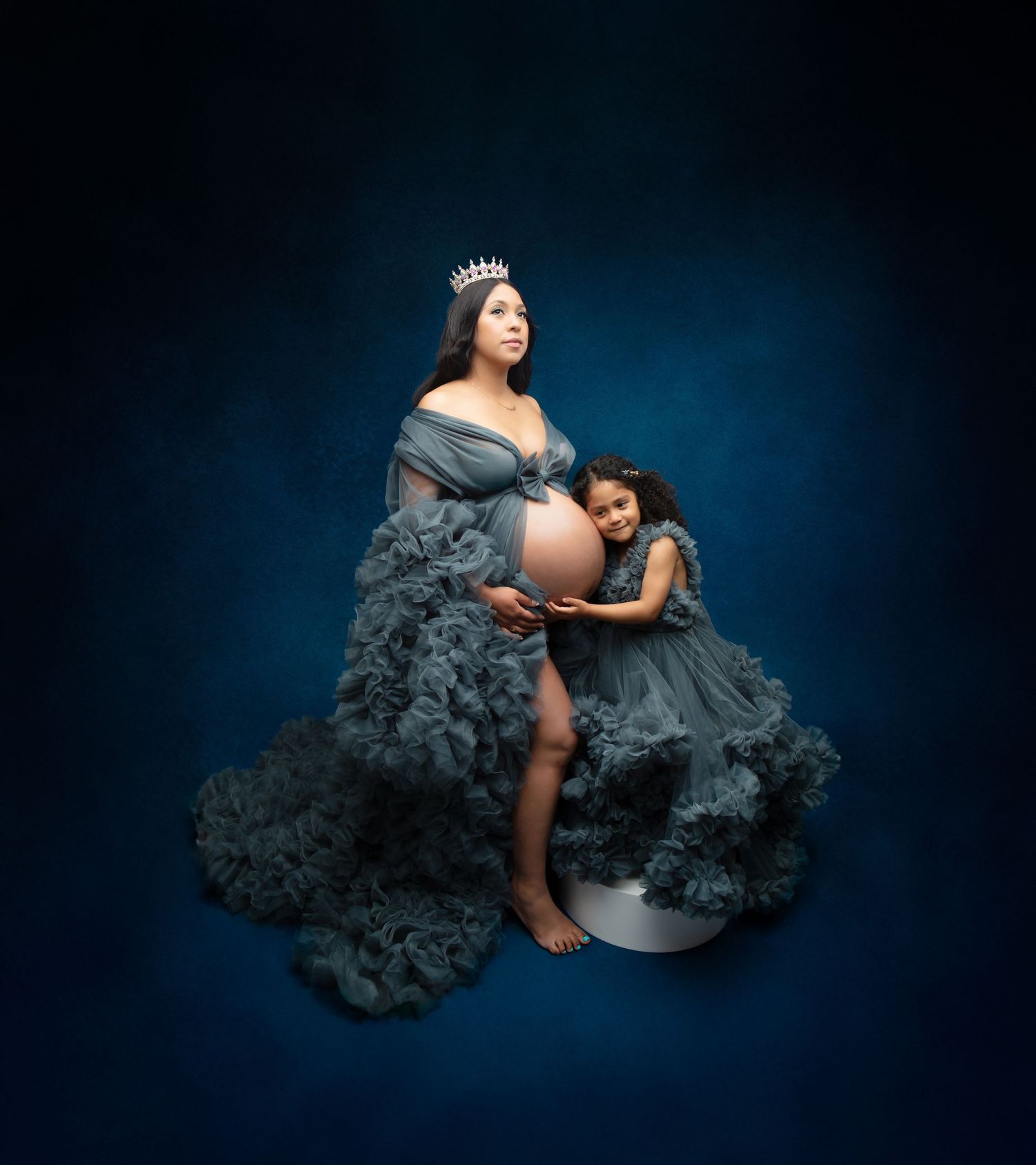 Pregnant woman in teal tulle gown with crown poses with young daughter in matching dress.