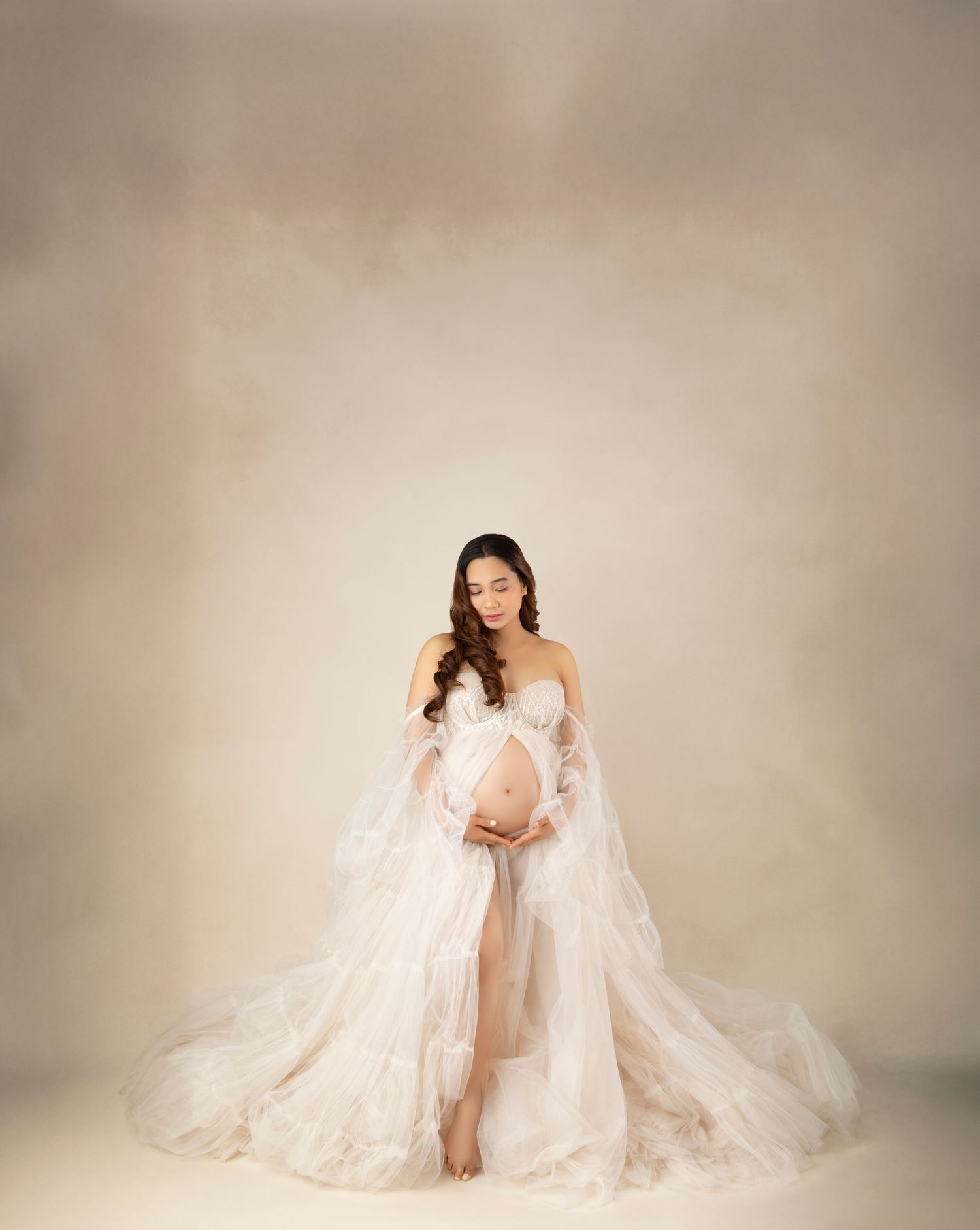 Pregnant woman in flowing white lace maternity gown posing gracefully in a soft beige studio.