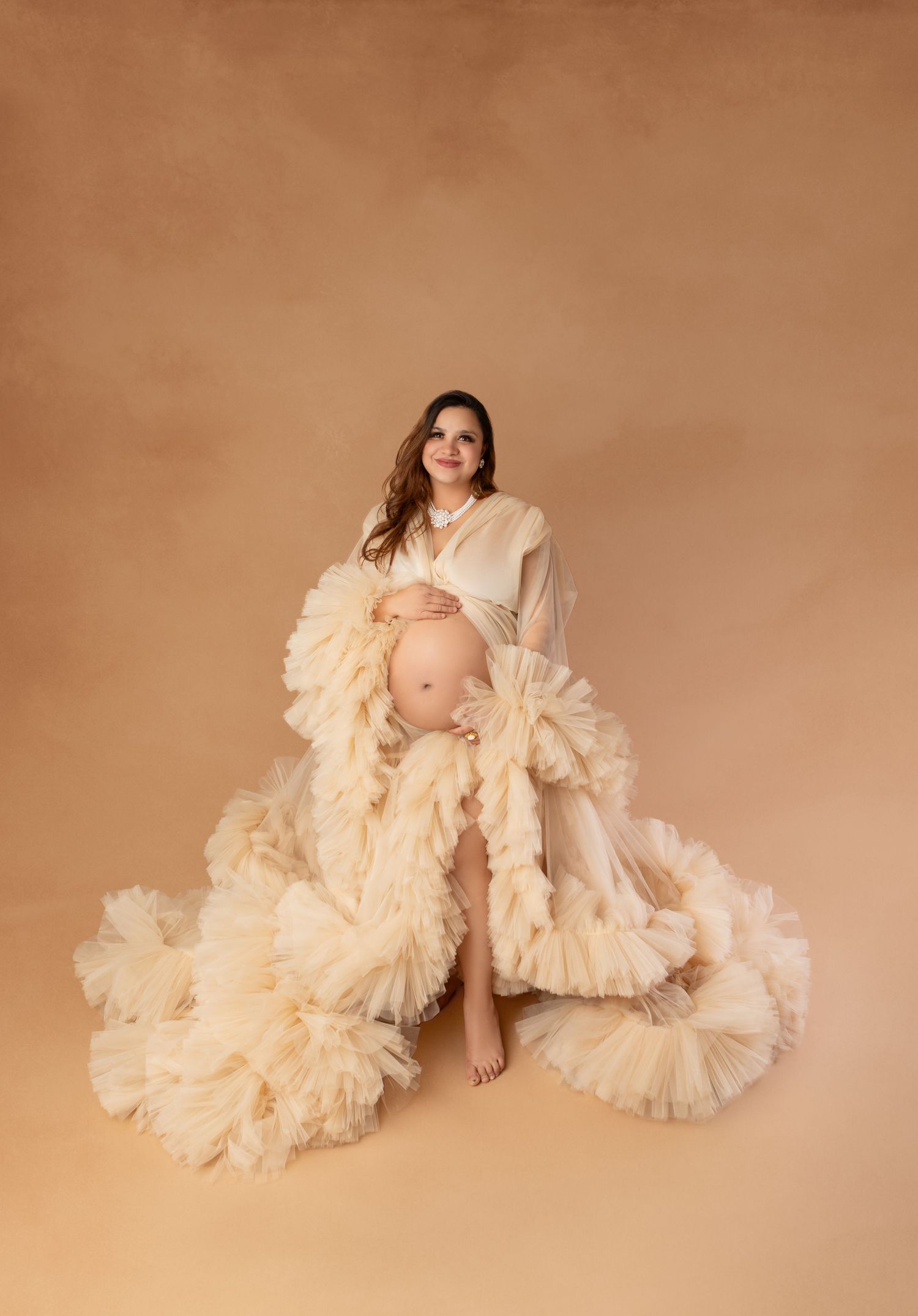Expecting mother wearing elegant cream ruffled tulle maternity dress posing in professional photo studio.