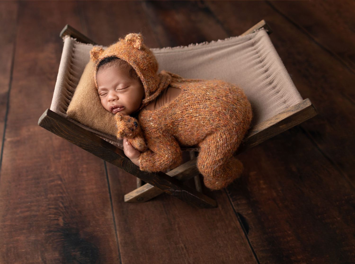 Newborn baby dressed in brown teddy bear outfit sleeping on rustic wooden cot.