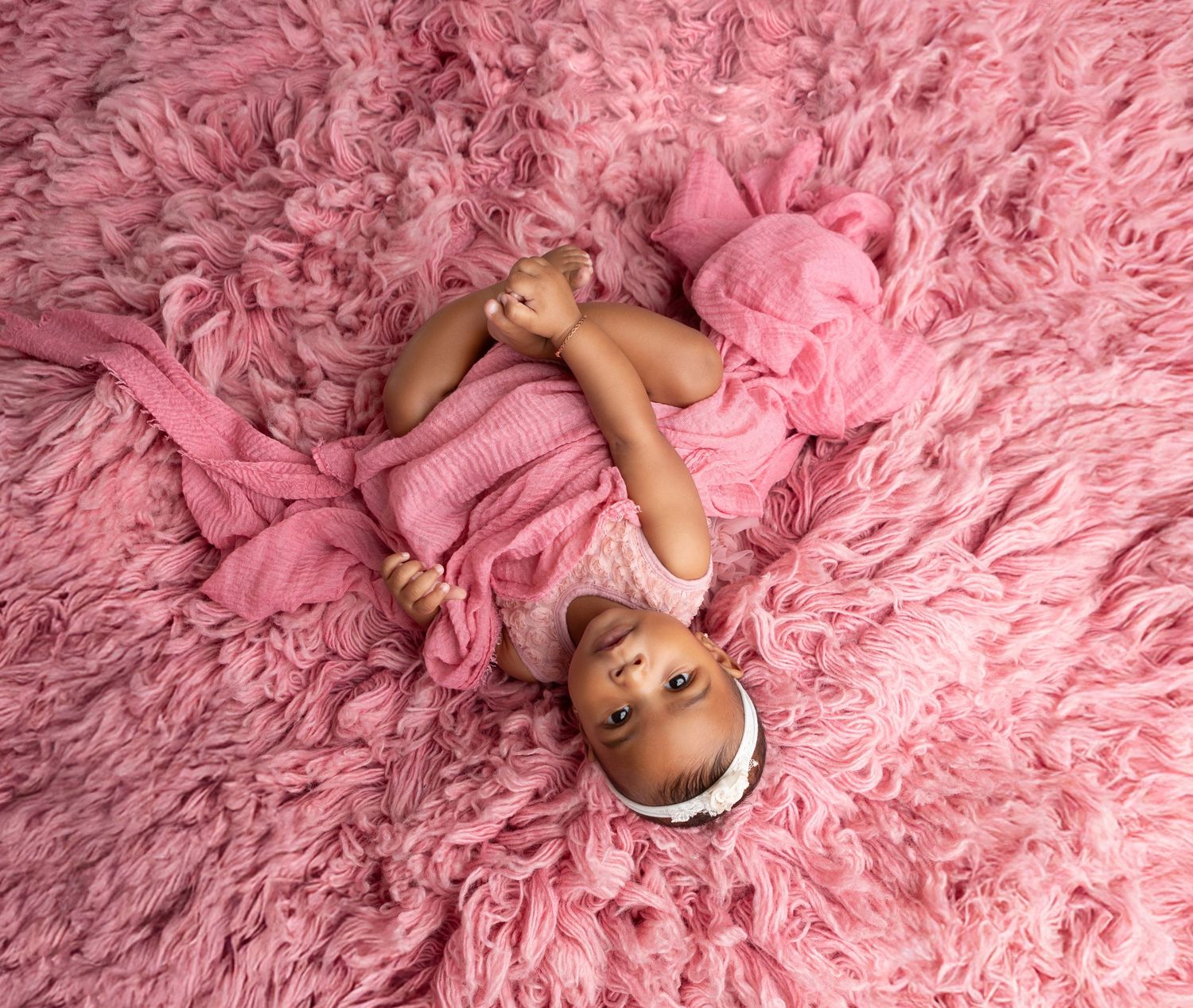 Newborn baby girl lying on pink fluffy rug wrapped in pink knit wrap with matching headband.