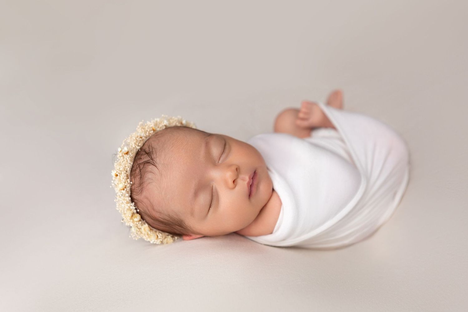 Sleeping newborn baby girl wrapped in white swaddle wearing a delicate floral crown headband on a neutral background.