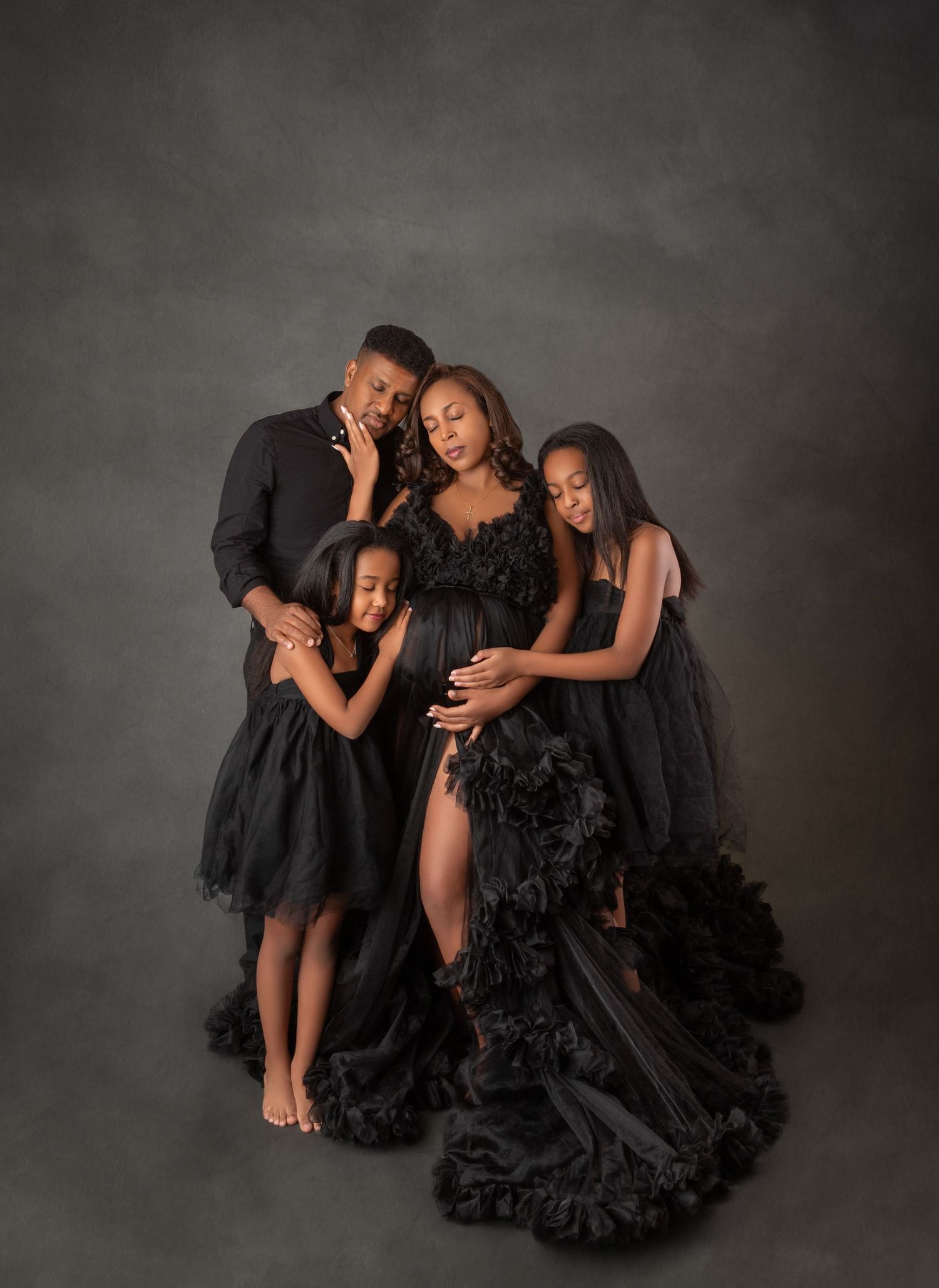 Family maternity photo session in matching black outfits with ruffled gown on gray backdrop.