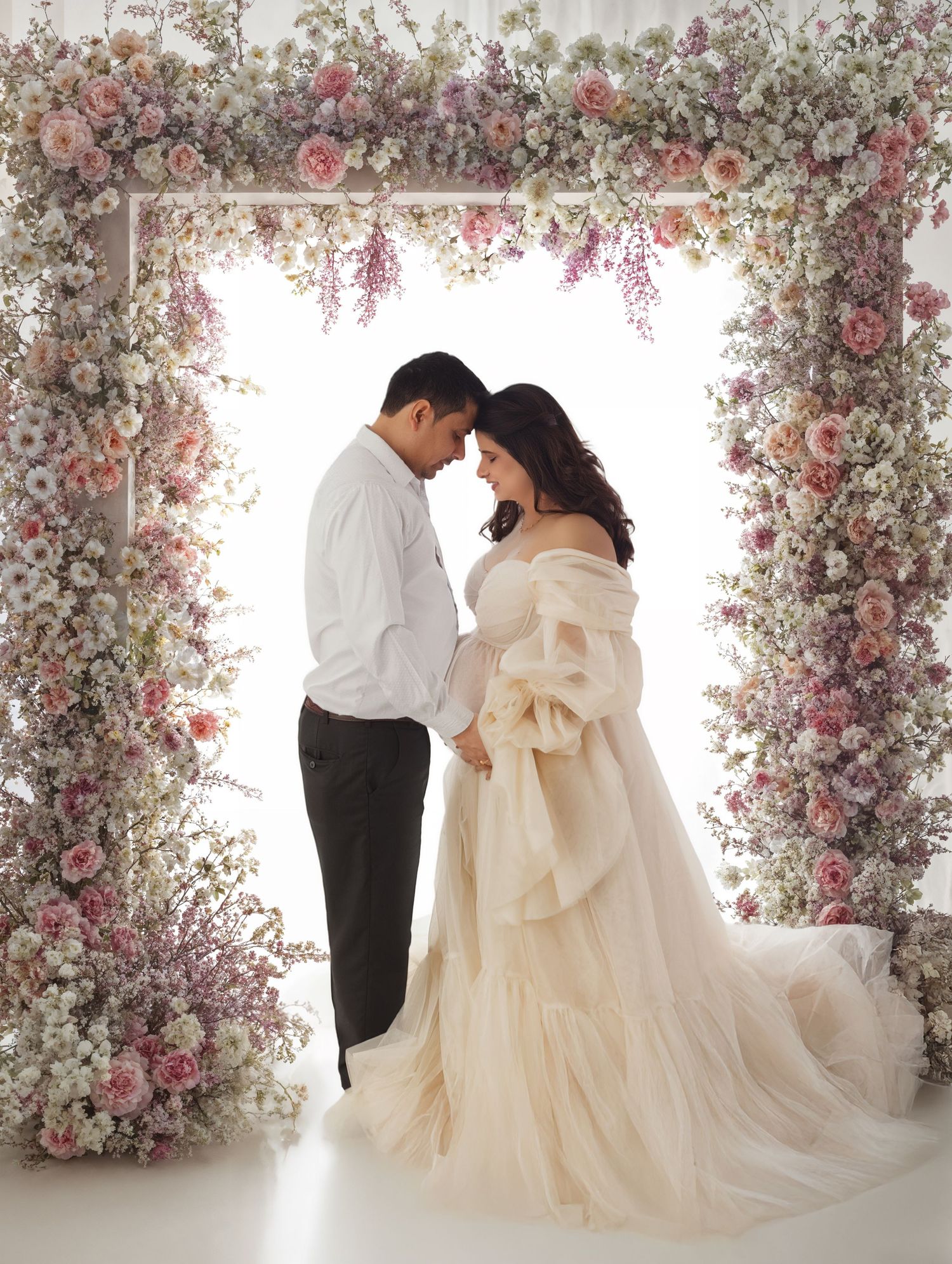 Couple maternity portrait under floral arch with pregnant woman in ivory off-shoulder gown.