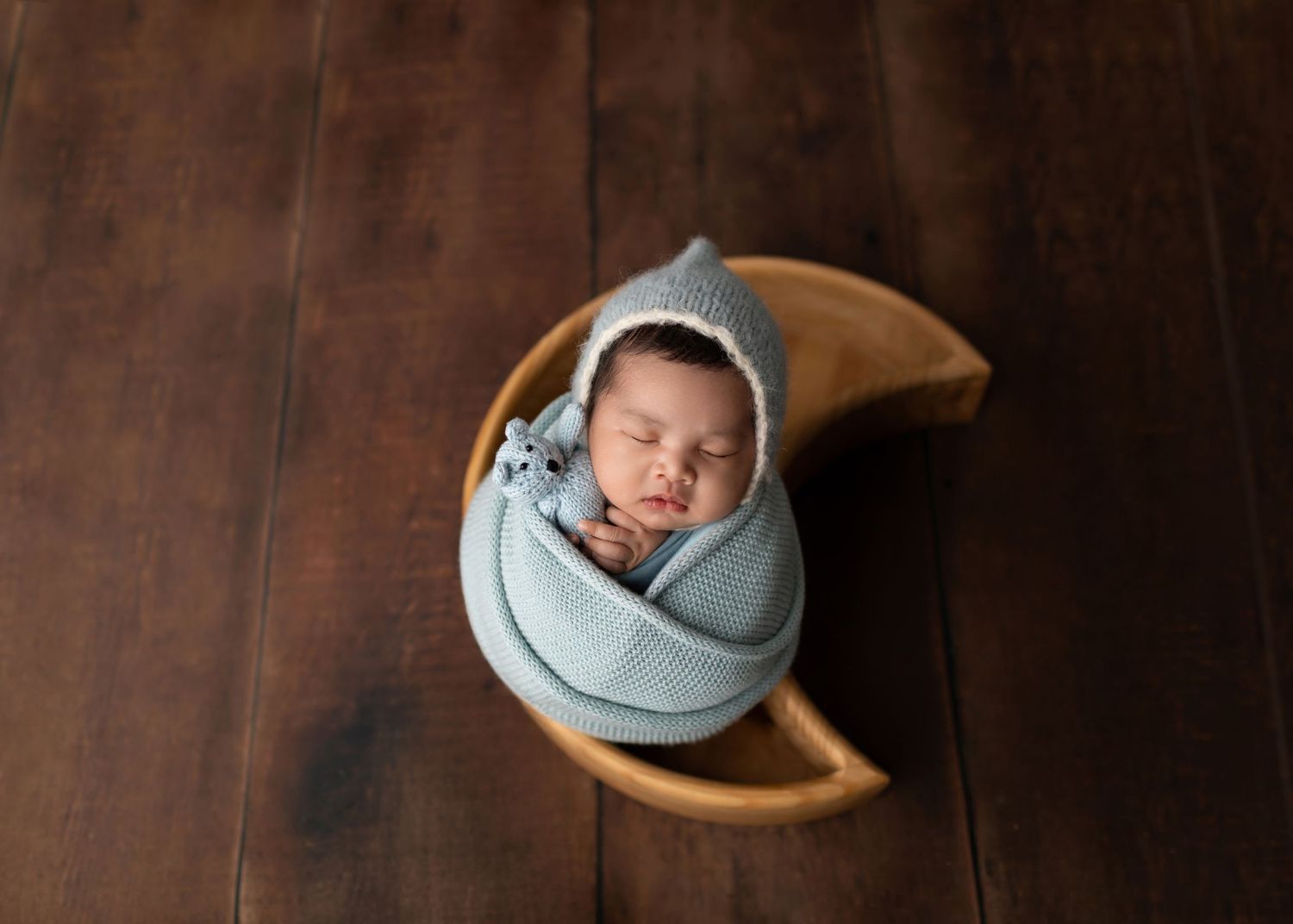 Swaddled newborn in blue wrap and gray bonnet sleeping in a wooden crescent moon bowl.