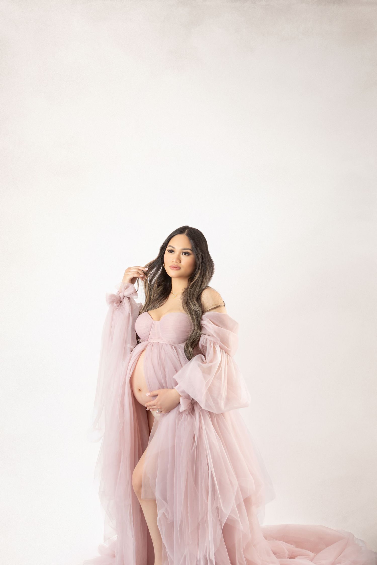 Pregnant woman in flowing pink tulle maternity gown posing gracefully in a bright white studio.