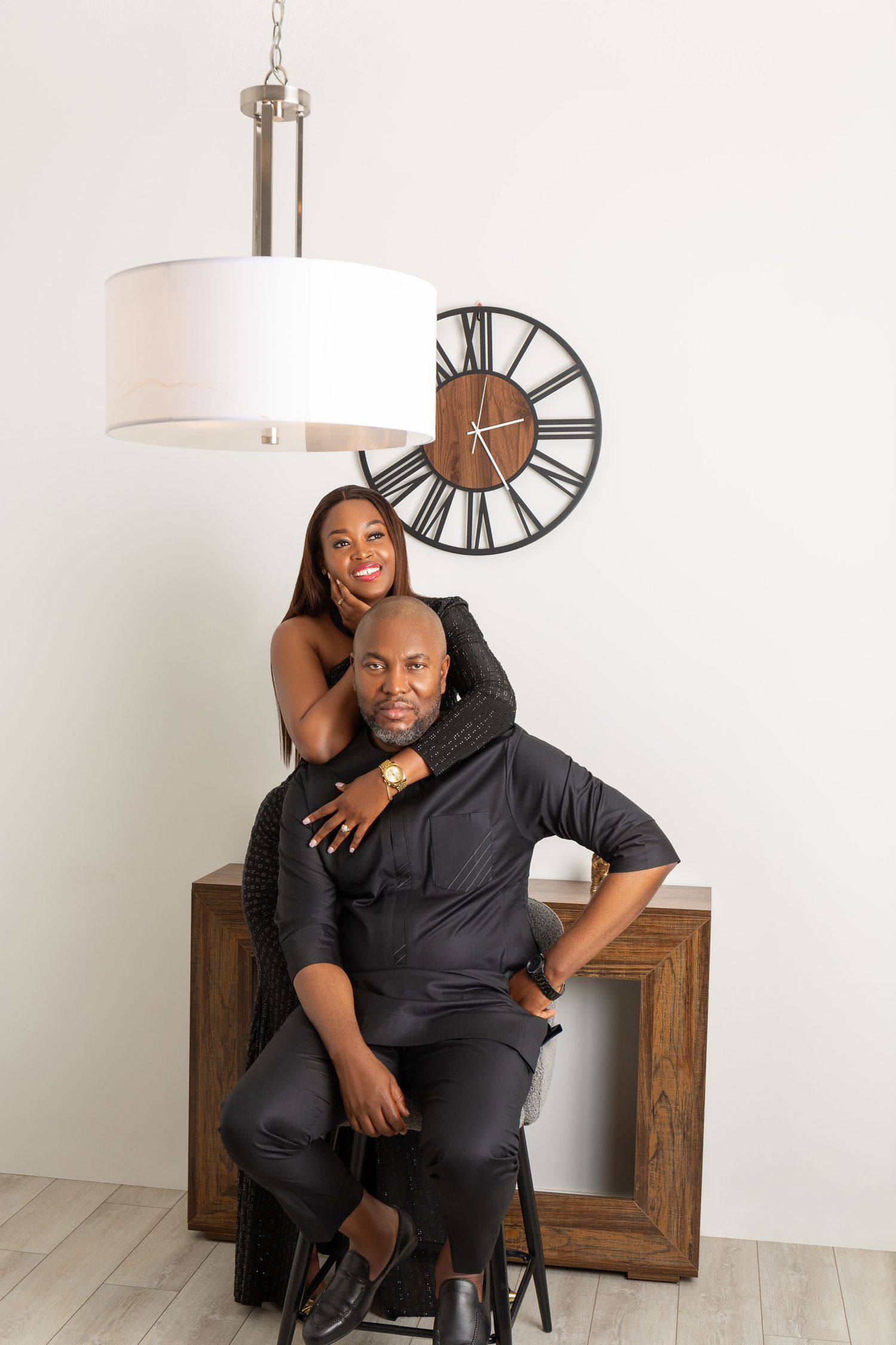 Elegant Black couple in matching black outfits pose together under a pendant light near a decorative clock.