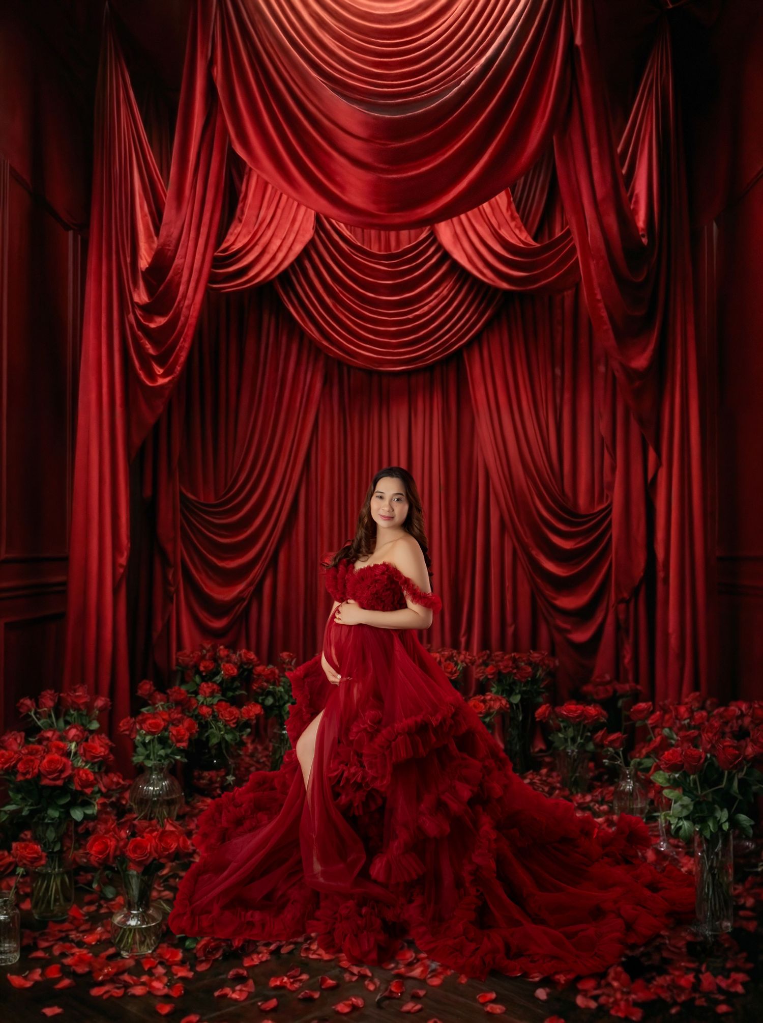 Pregnant woman in red ruffled gown poses among roses before dramatic red velvet curtains in elegant maternity portrait.