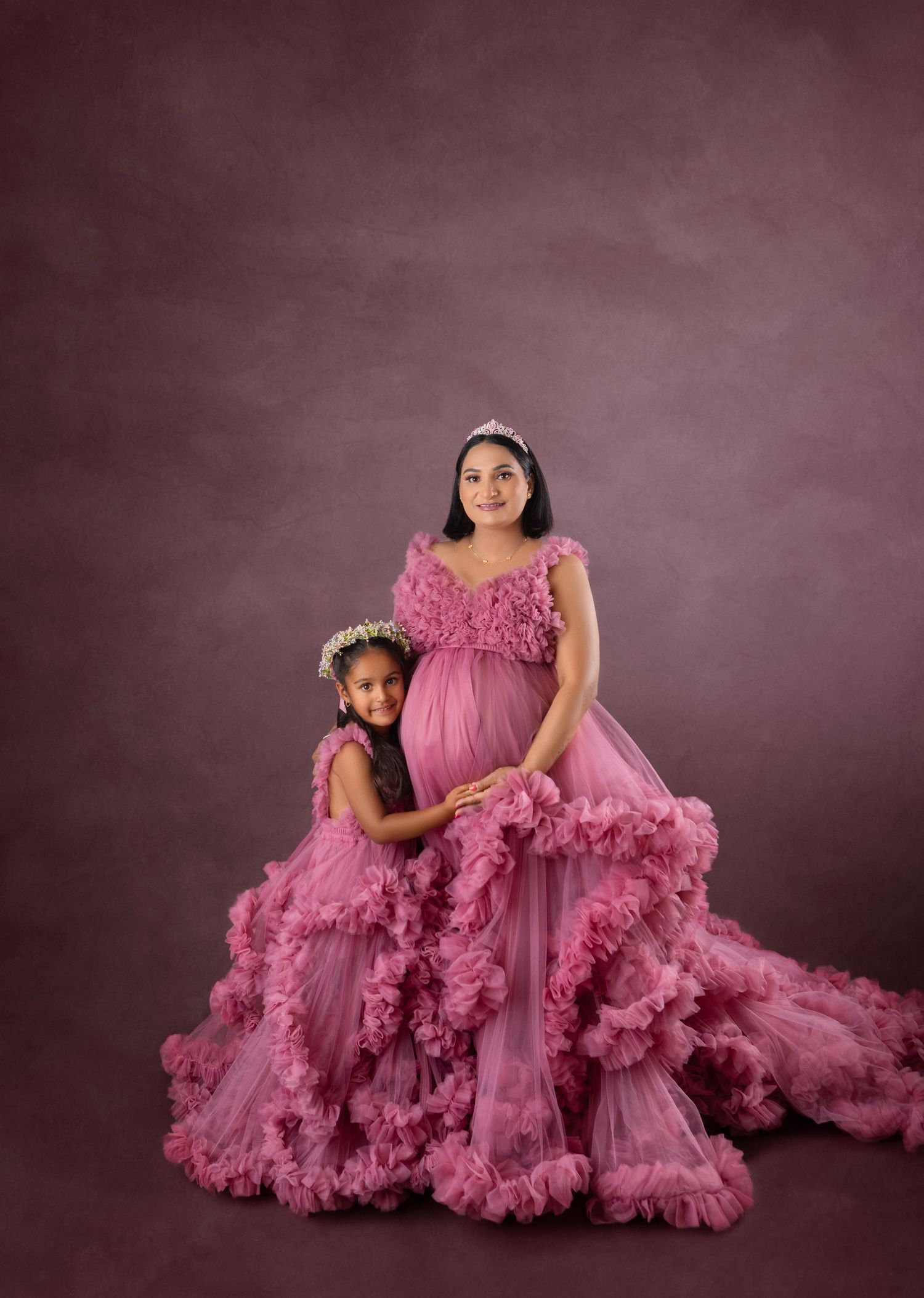 Pregnant woman and young girl in matching pink ruffled gowns pose for a maternity portrait session.