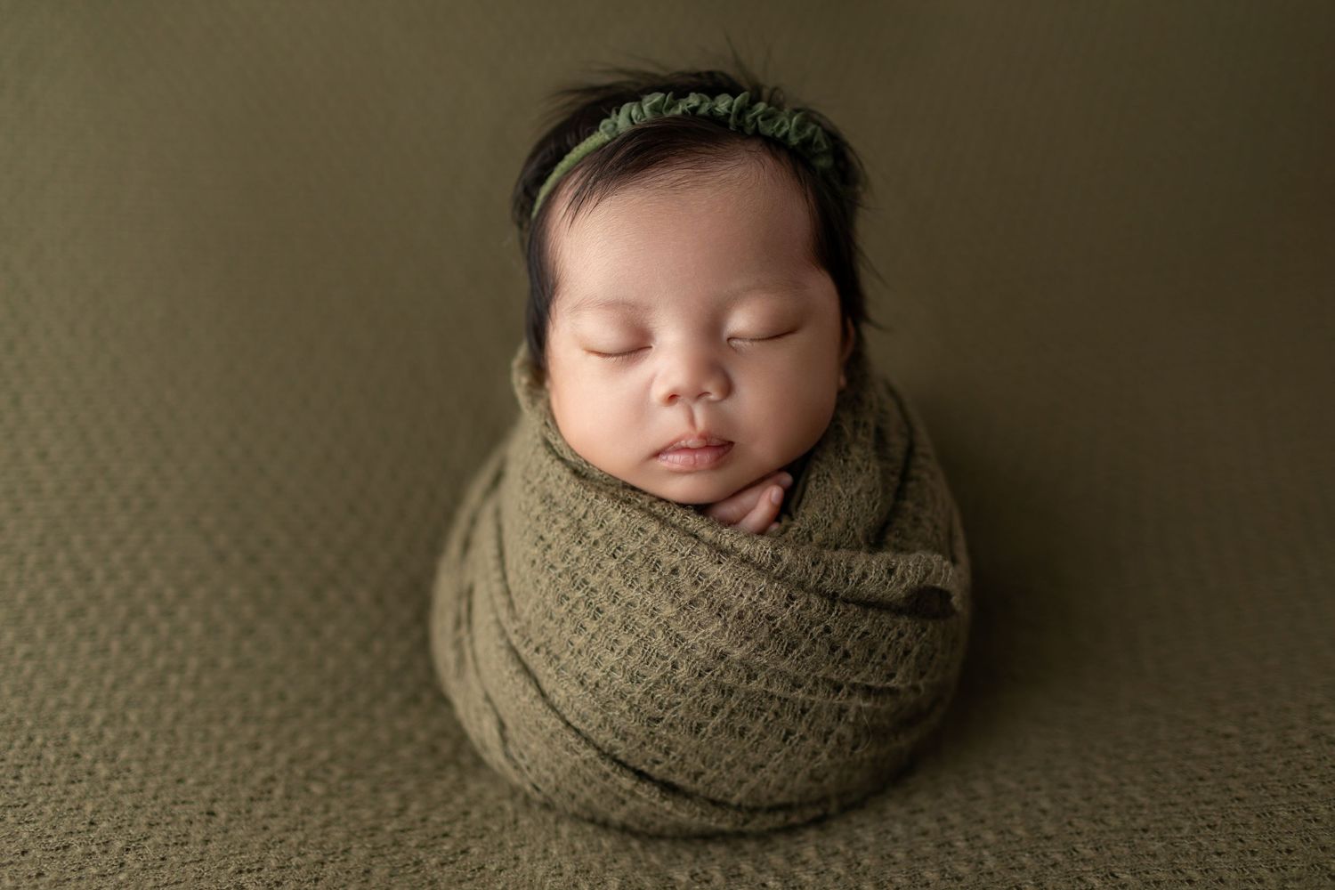 Newborn baby wrapped snugly in olive green swaddle with matching headband on green textured backdrop.