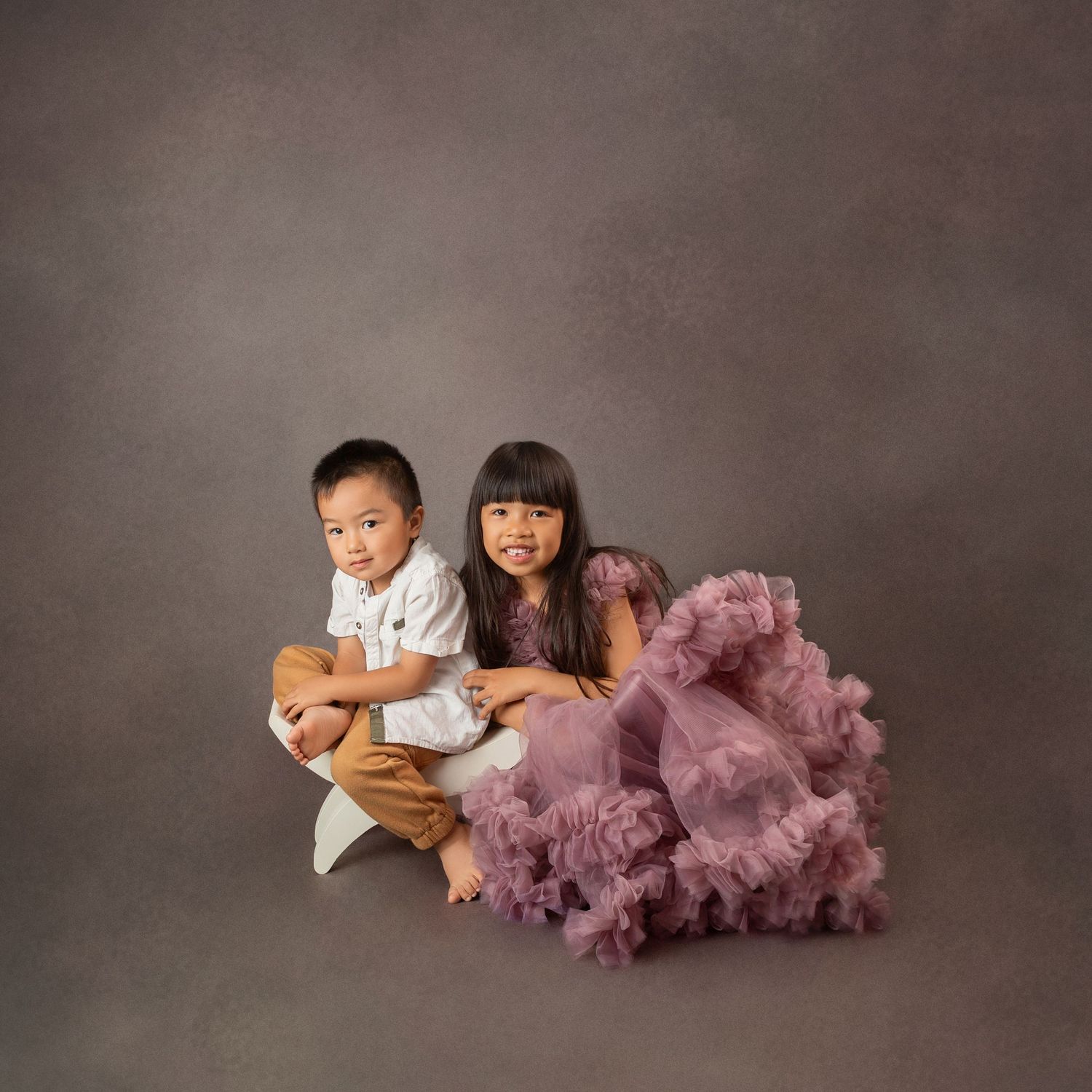 Two young children pose together in a studio, a boy in white shirt and a girl in a purple tulle dress.