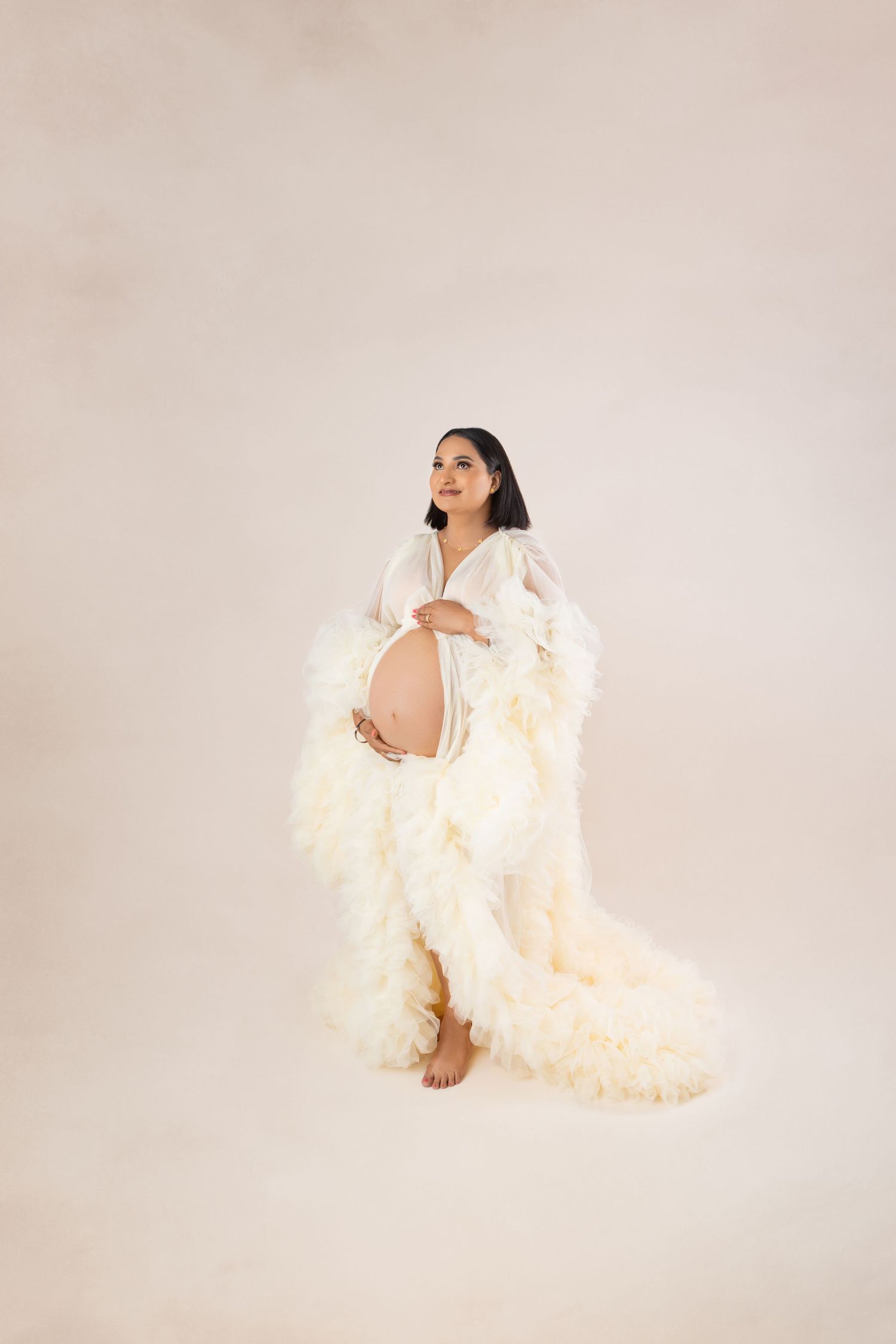 Pregnant woman in elegant white ruffled maternity gown cradling baby bump in a professional studio photo.