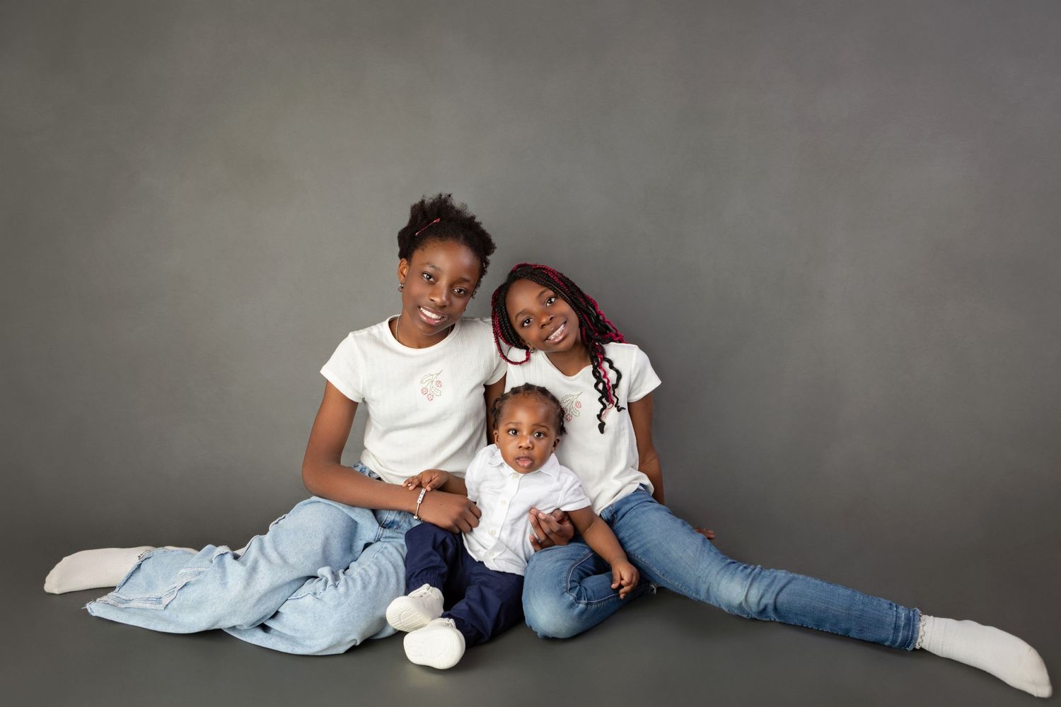 Two Black sisters sit on the floor with a baby between them in a cozy studio family photo.