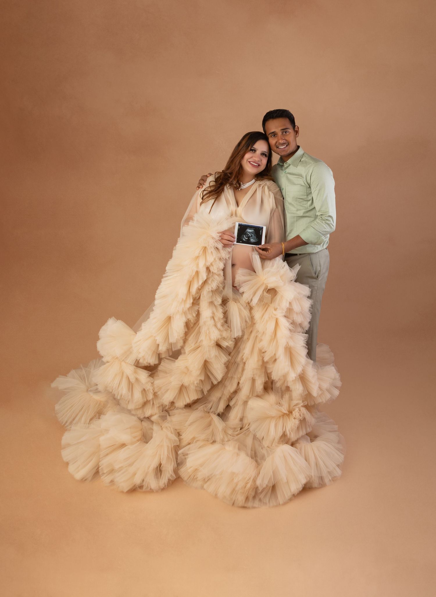 Couple holding ultrasound photo together, woman wearing cream ruffled tulle maternity gown in studio.