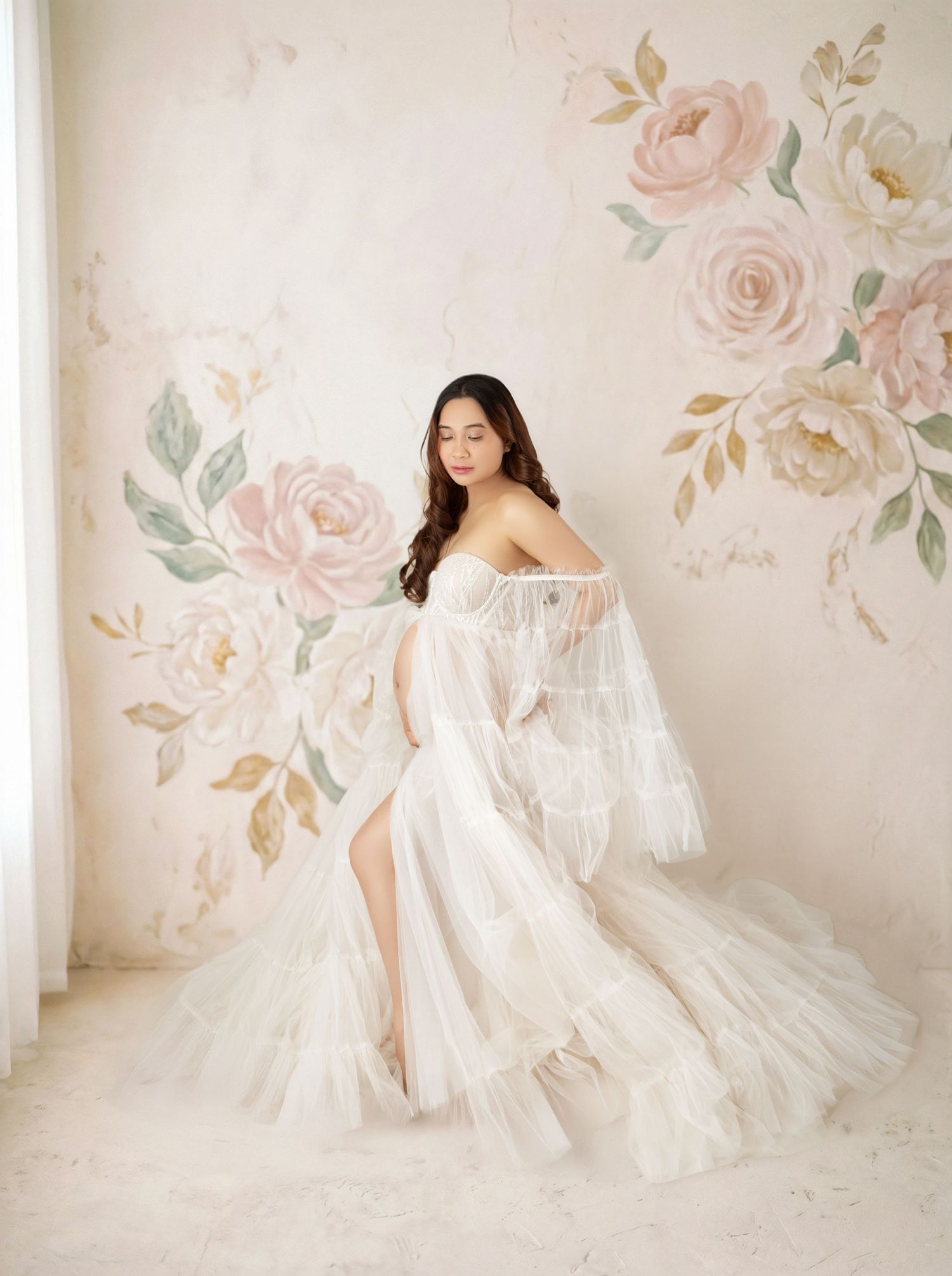 Woman in white tulle off-shoulder gown poses gracefully against a floral painted backdrop.