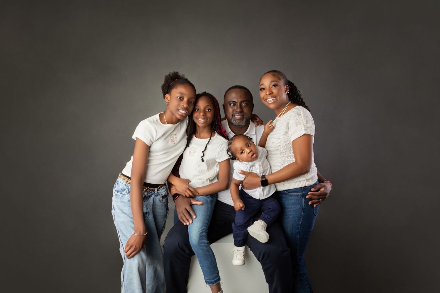 Happy Black family of five posing together in matching white shirts and jeans against gray background.
