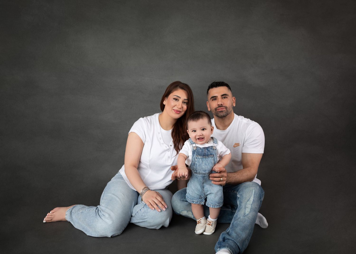 Smiling family portrait with baby in denim overalls against grey studio background.