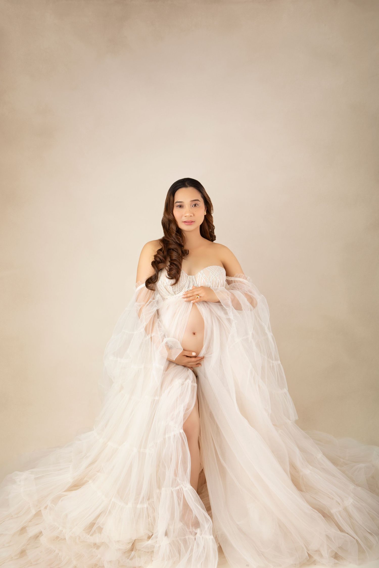 A pregnant woman in a flowing white tulle maternity gown cradles her belly on a beige background.