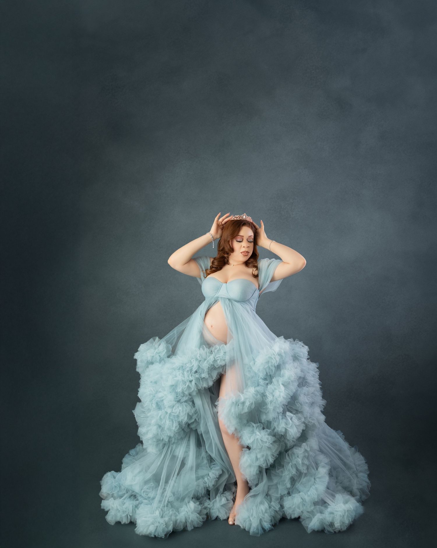 Pregnant woman in dramatic ruffled teal tulle gown posing against a dark gray studio background.