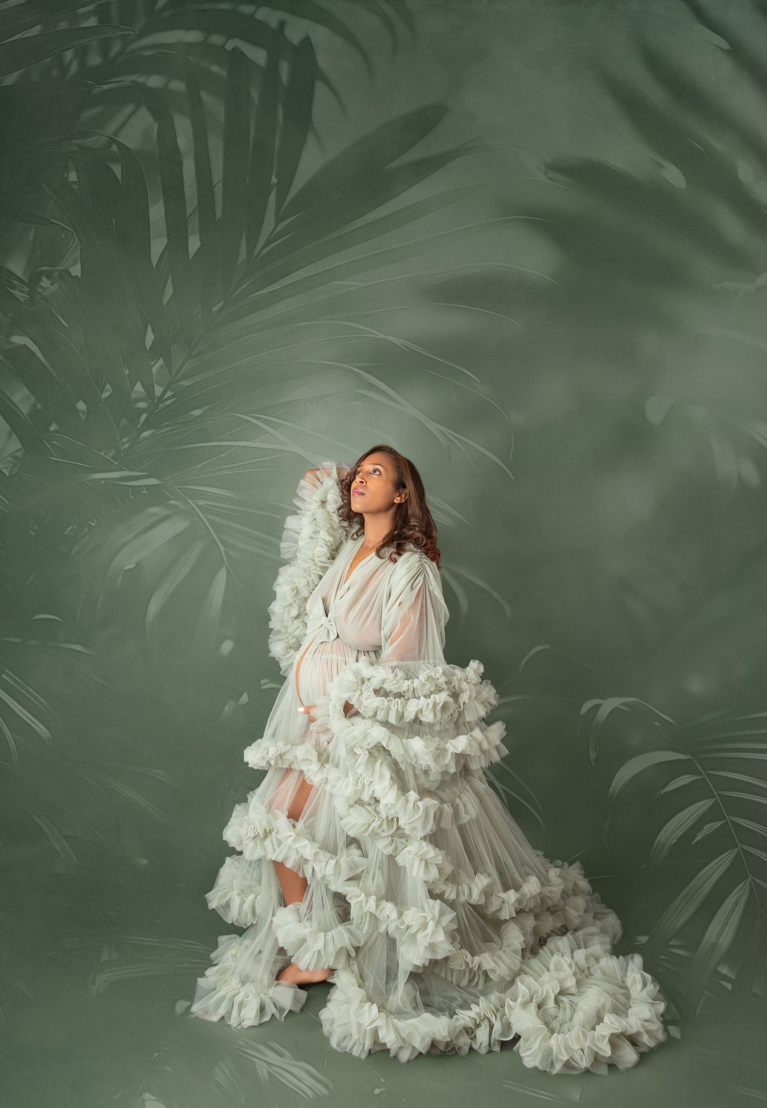 Pregnant woman in white ruffled gown looks upward dramatically against a green palm leaf studio backdrop.