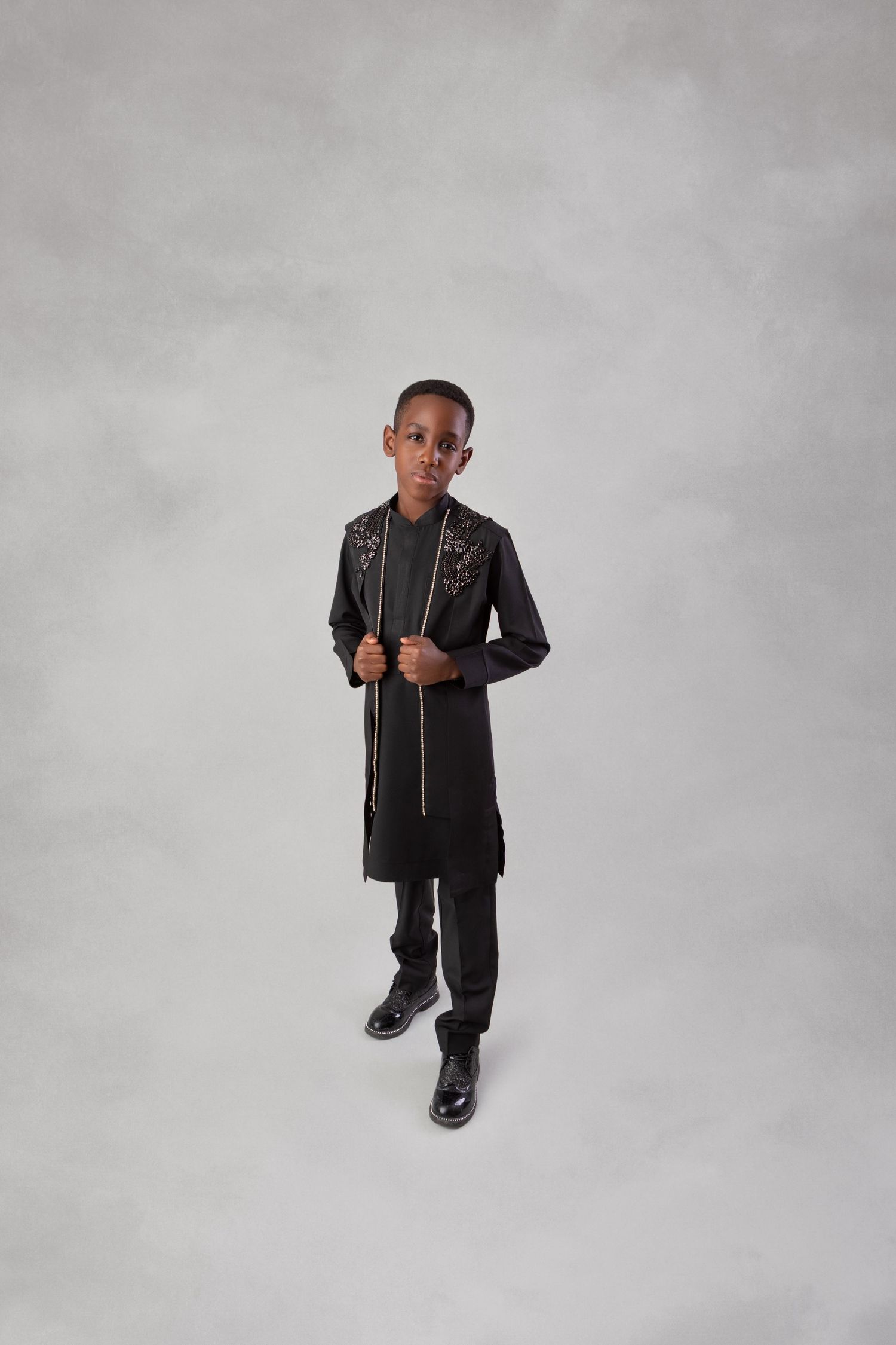 Young boy in elegant black embroidered long jacket with zipper detail and dress pants posing in studio.