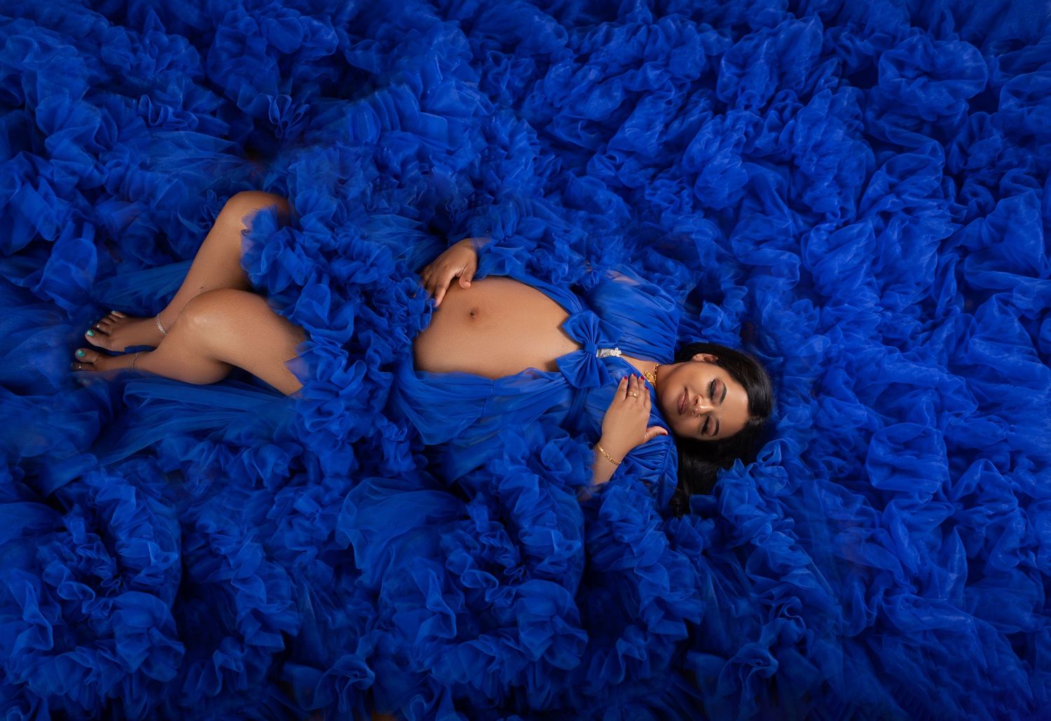 Pregnant woman lying in a flowing royal blue tulle gown spread across the floor in a dramatic maternity photo.