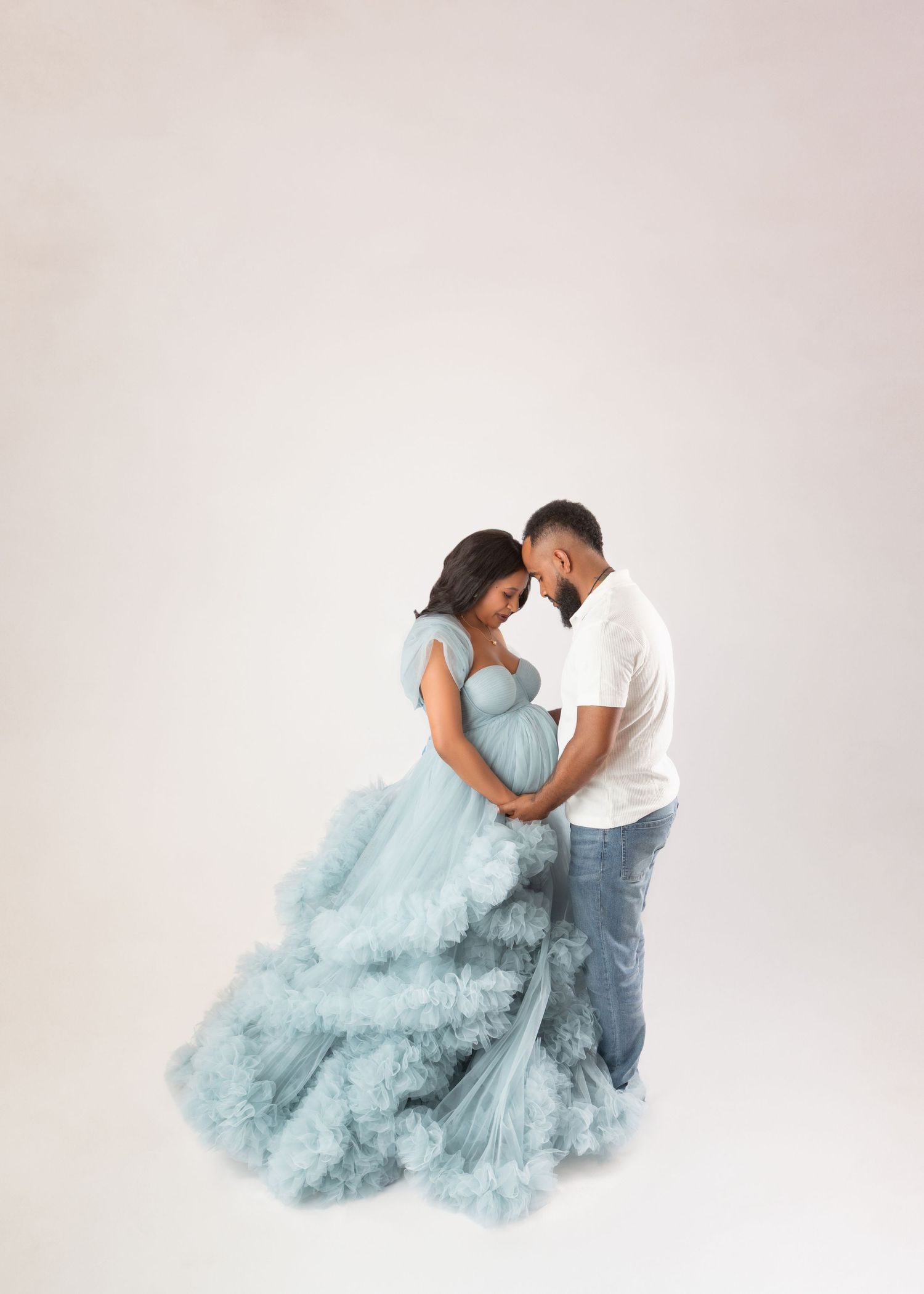 Pregnant couple in studio, woman in flowing blue tulle gown, man in white shirt and jeans, foreheads touching tenderly.