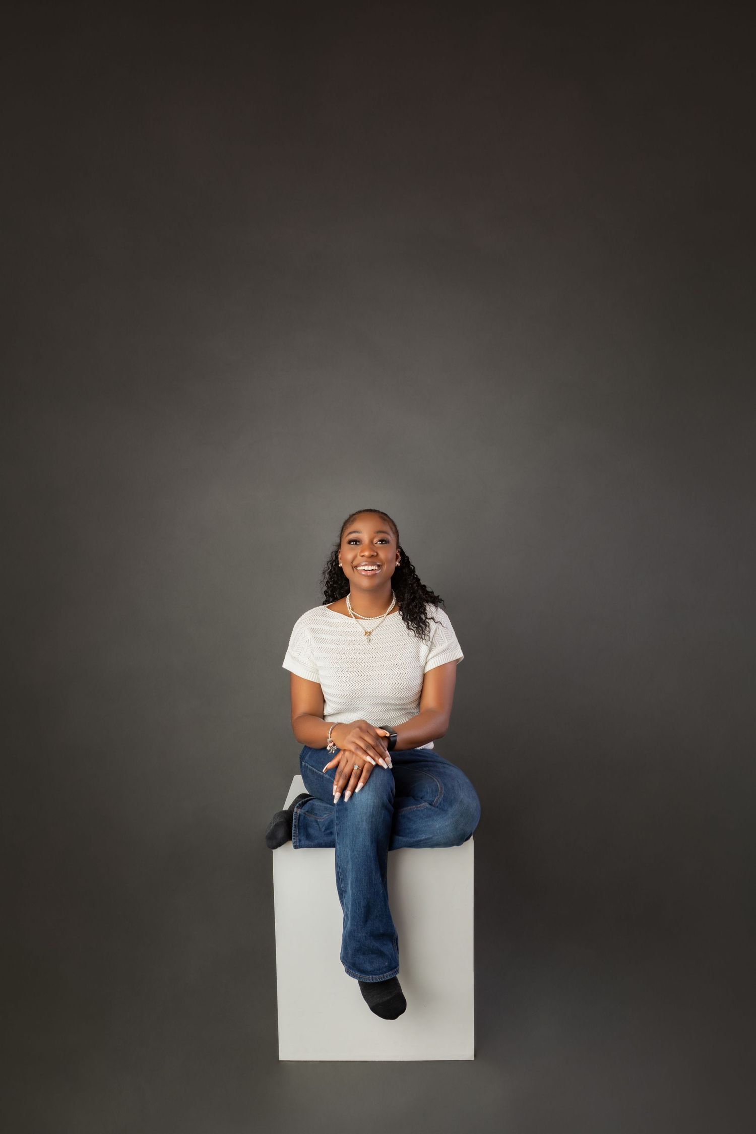 Young Black woman sitting on white cube, wearing white tee and jeans, smiling against gray background.