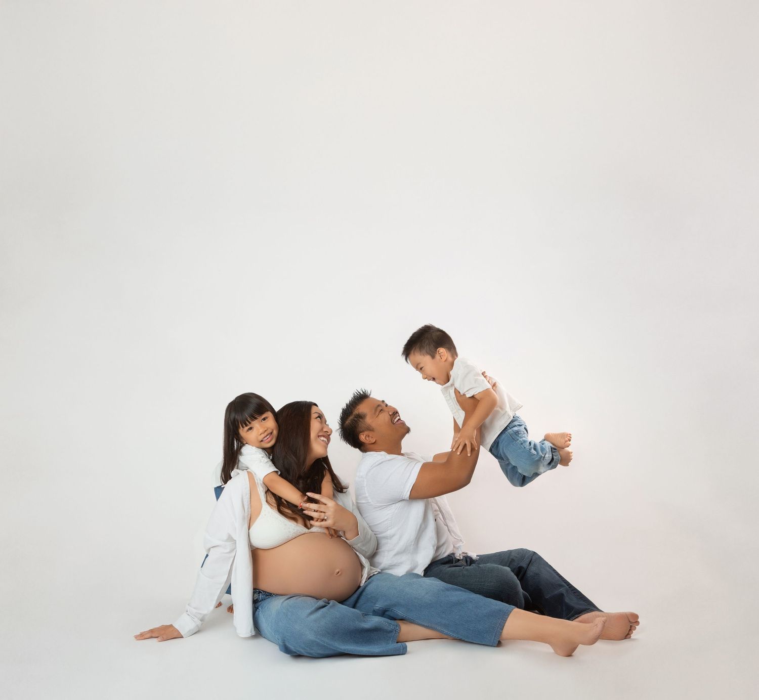 Pregnant woman sitting on floor with husband and two young children, wearing white tops and jeans, on white background.