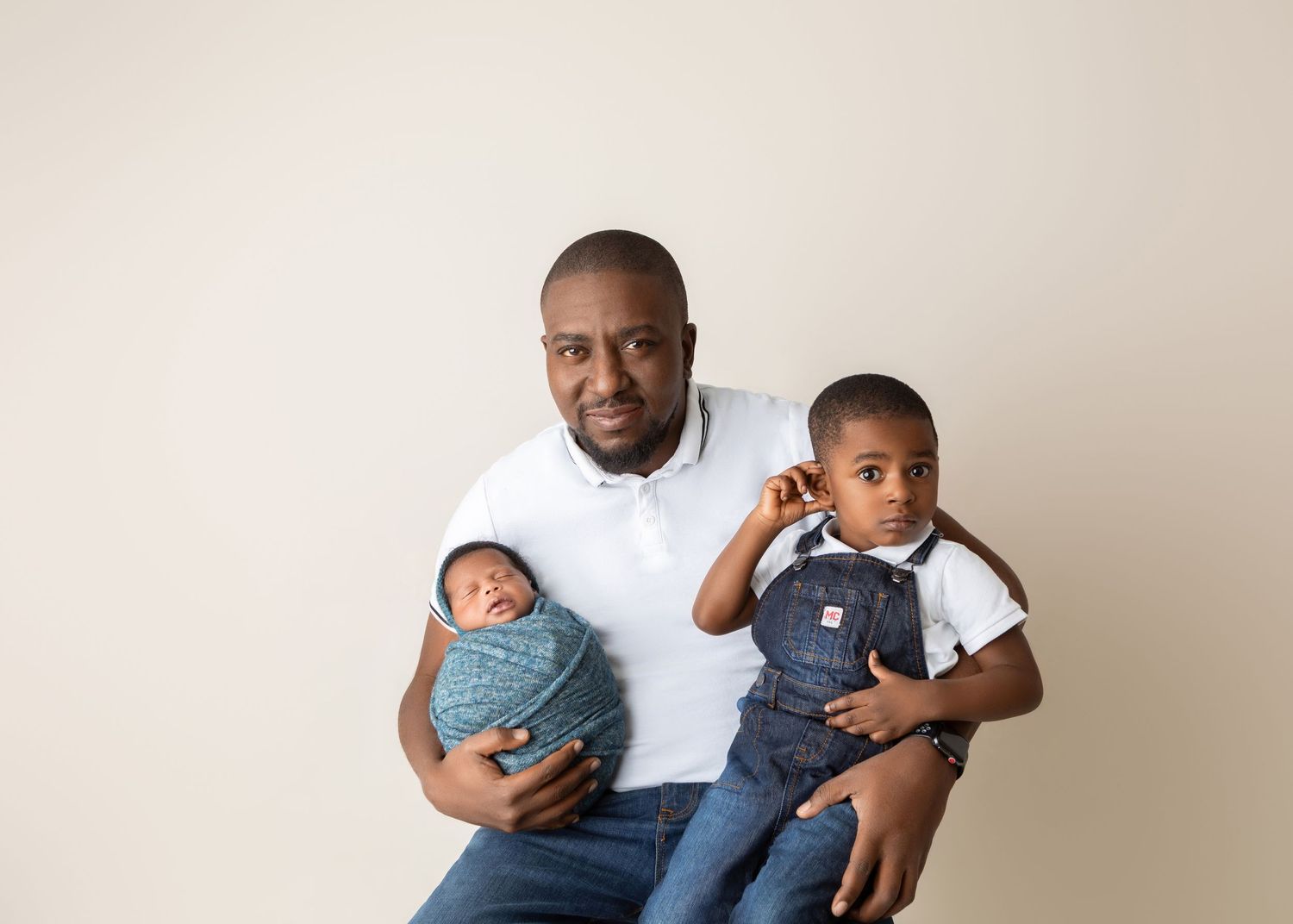 Father holding newborn wrapped in blue and toddler son in denim overalls.