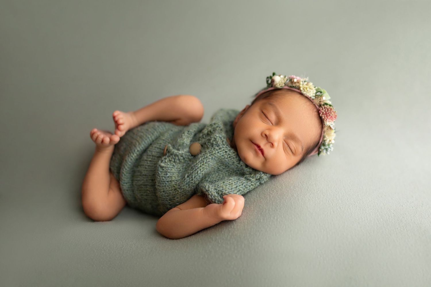 Sleeping newborn baby in green knit romper and floral headband on sage green background.