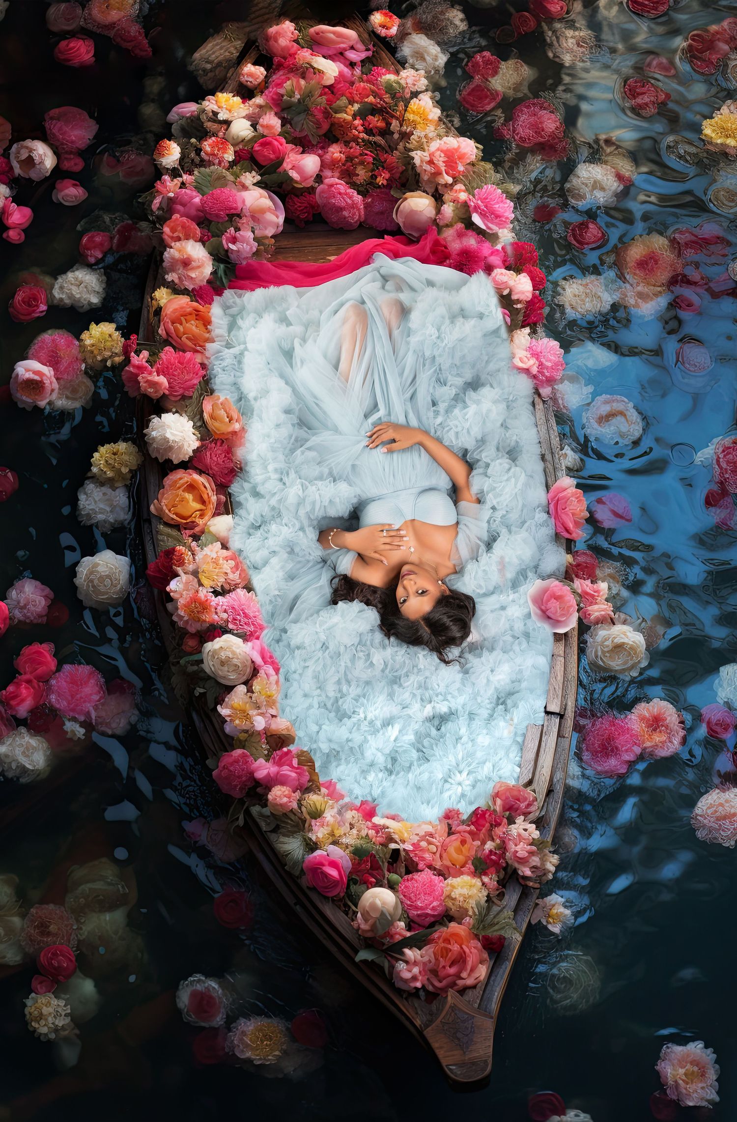 A pregnant woman in white lies in a flower-filled wooden boat floating on water in artistic portrait.