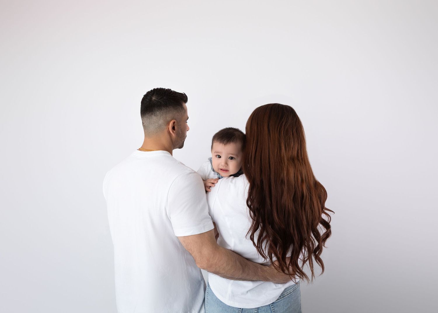 Parents in white tees hold curious baby facing white studio wall, back view.