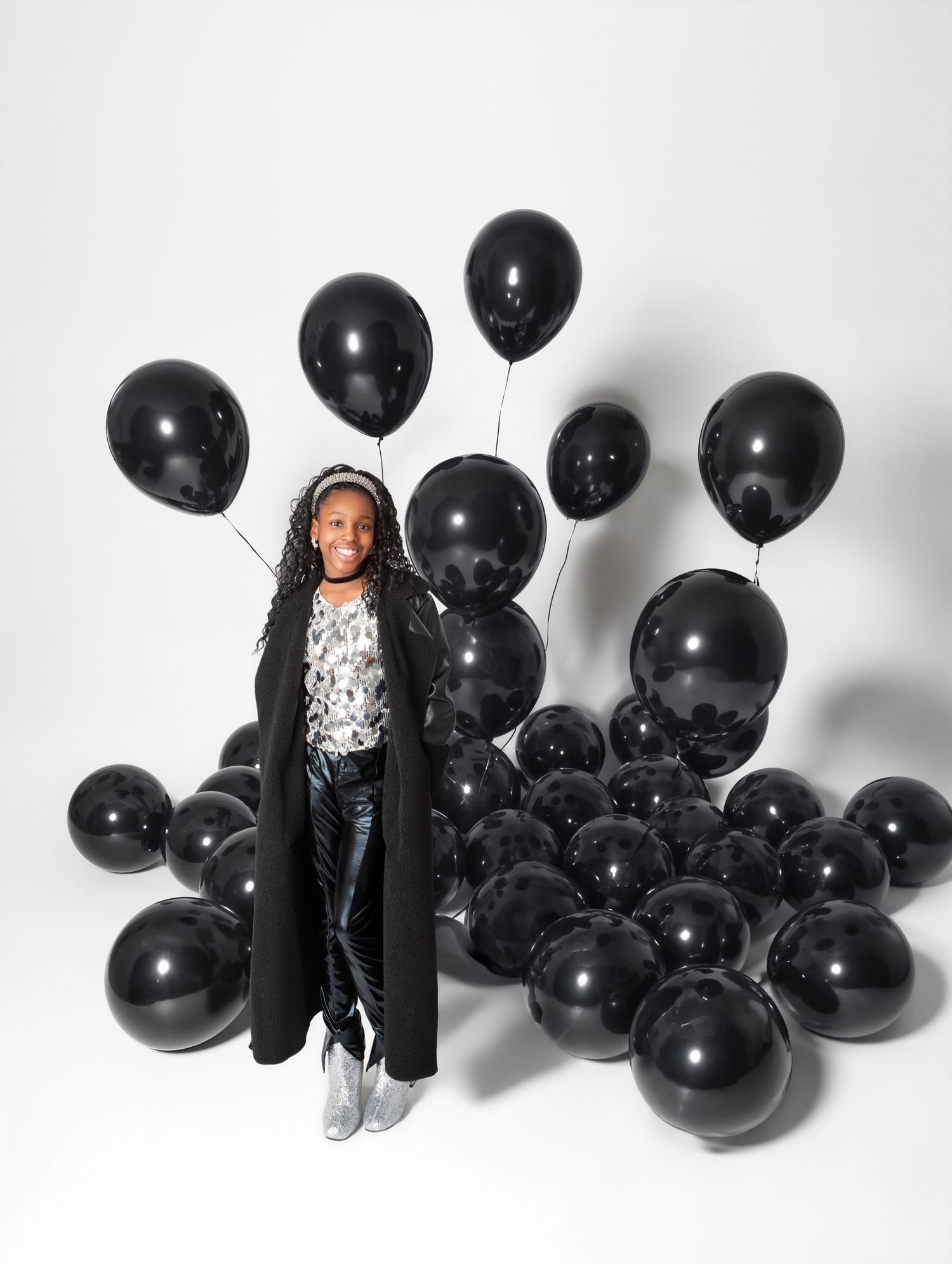 Young girl in black coat smiling among large pile of black balloons in white studio.