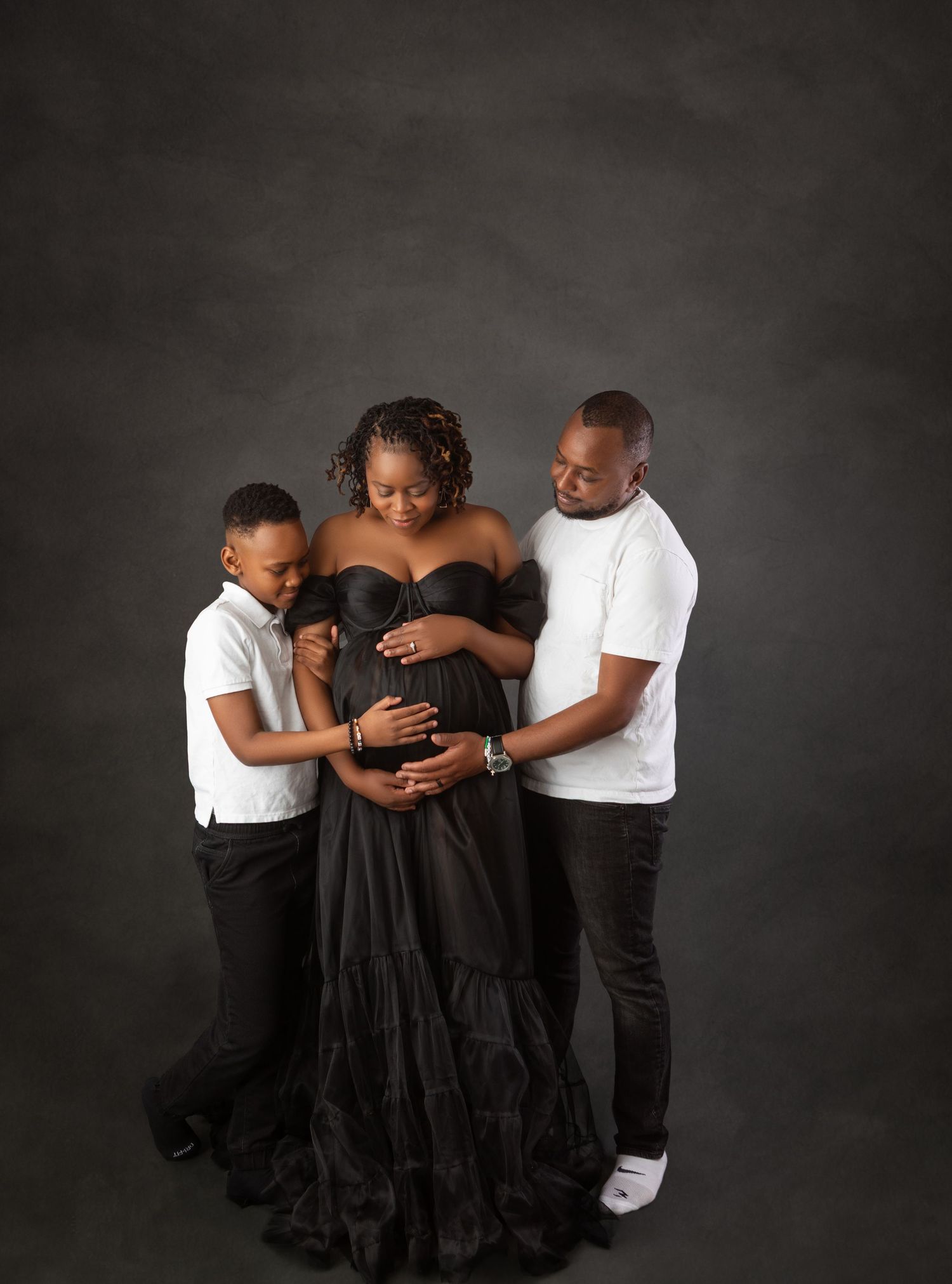 Family maternity portrait with pregnant woman in black gown flanked by partner and child on gray backdrop.