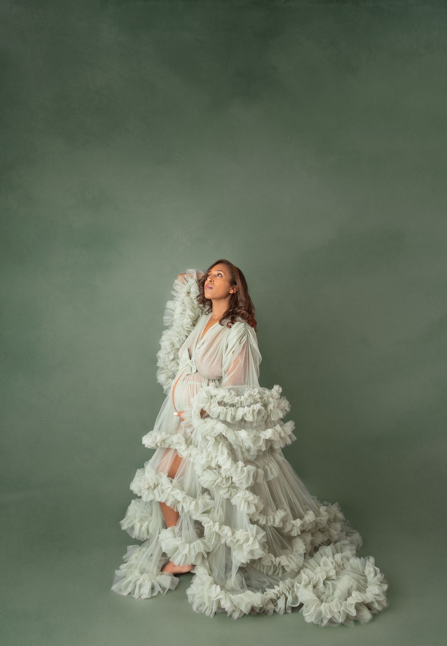Pregnant woman in white ruffled tulle gown poses gracefully against sage green studio backdrop.