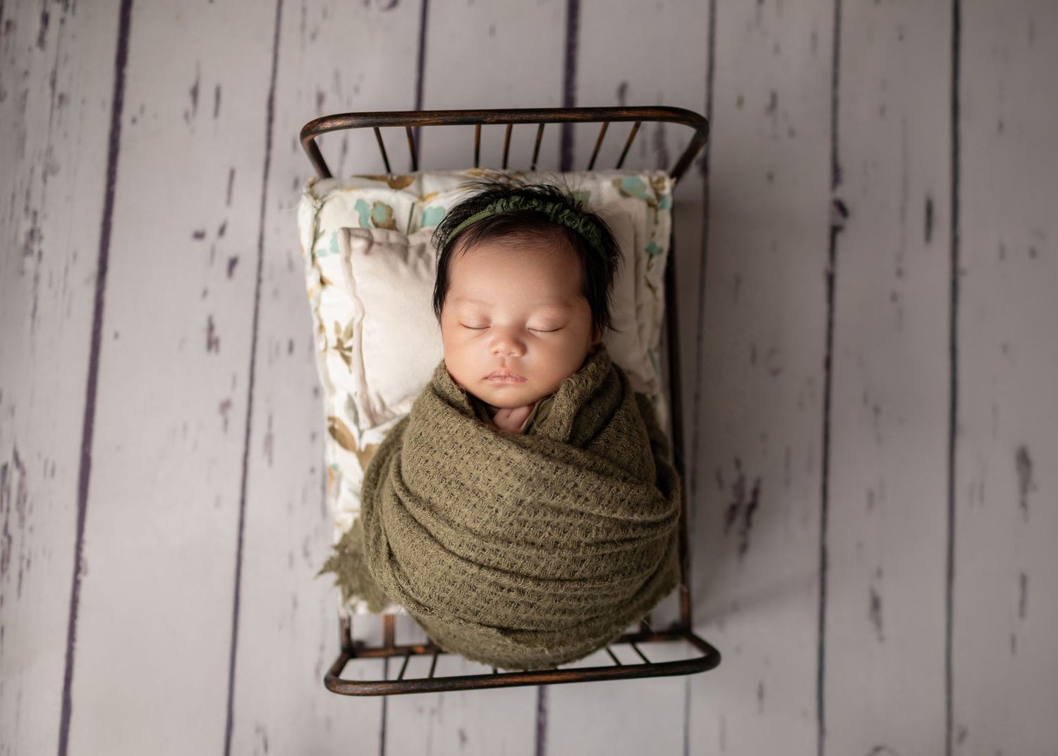 Newborn baby swaddled in olive green wrap sleeping peacefully in a vintage metal crib for a professional photo.