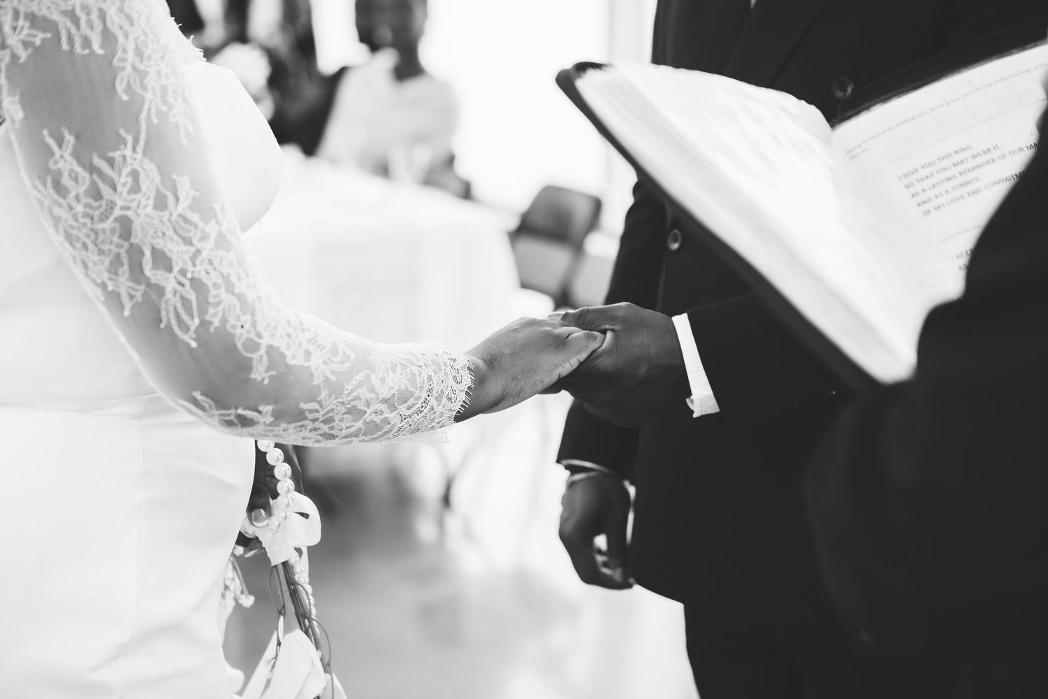 Black and white wedding ceremony photo of couple exchanging vows with officiant reading from book.