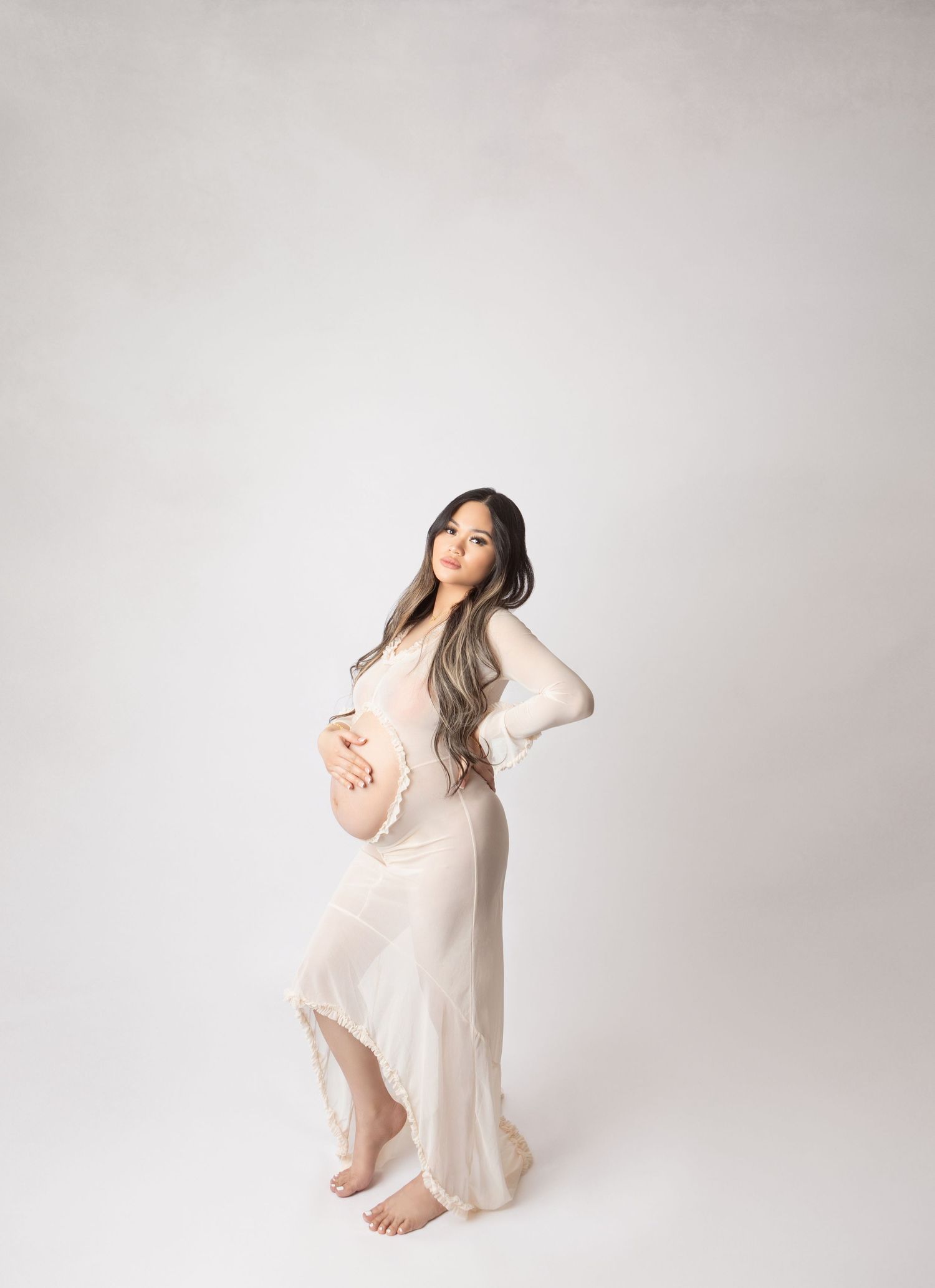 Expectant mother in sheer ivory dress cradling baby bump while posing in a minimalist white studio.
