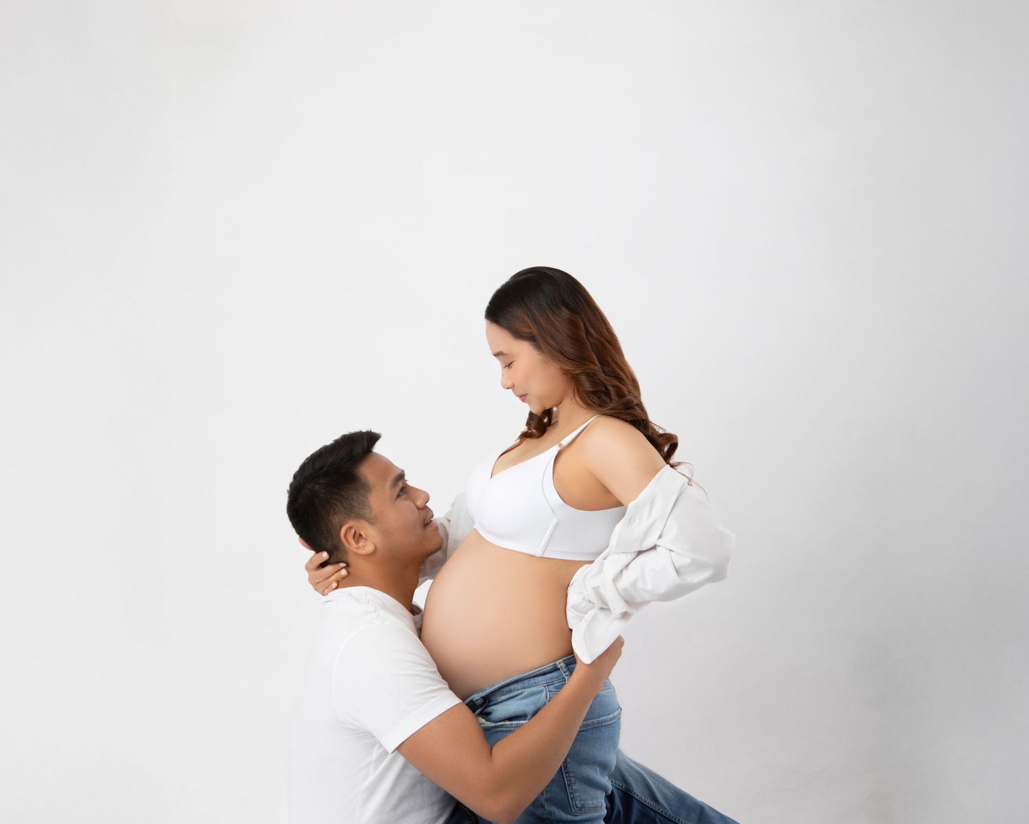 Man kneeling gazes up lovingly at pregnant partner in white bra top and open shirt on white backdrop.