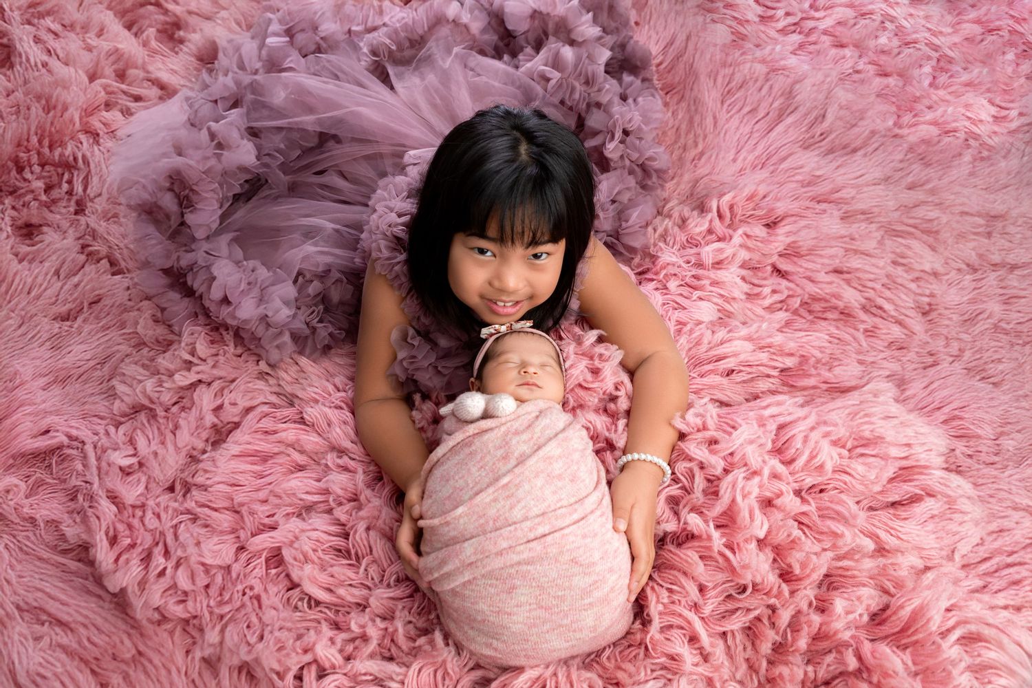 Young girl in purple dress tenderly holds swaddled newborn baby on pink fluffy backdrop.