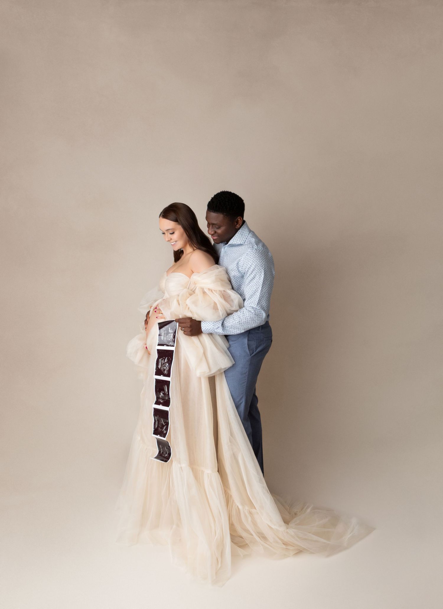 Expecting couple holds ultrasound photos while embracing in neutral-toned studio maternity portrait session.