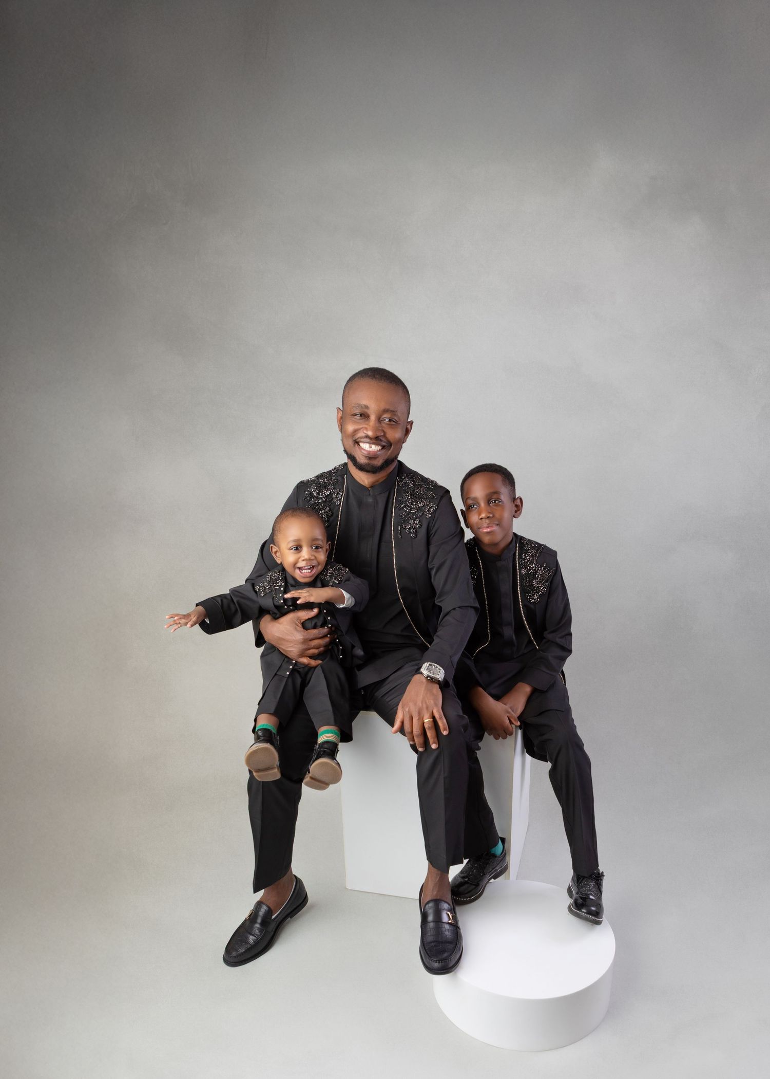 Father in black embroidered suit poses with two sons in matching outfits on white cube.